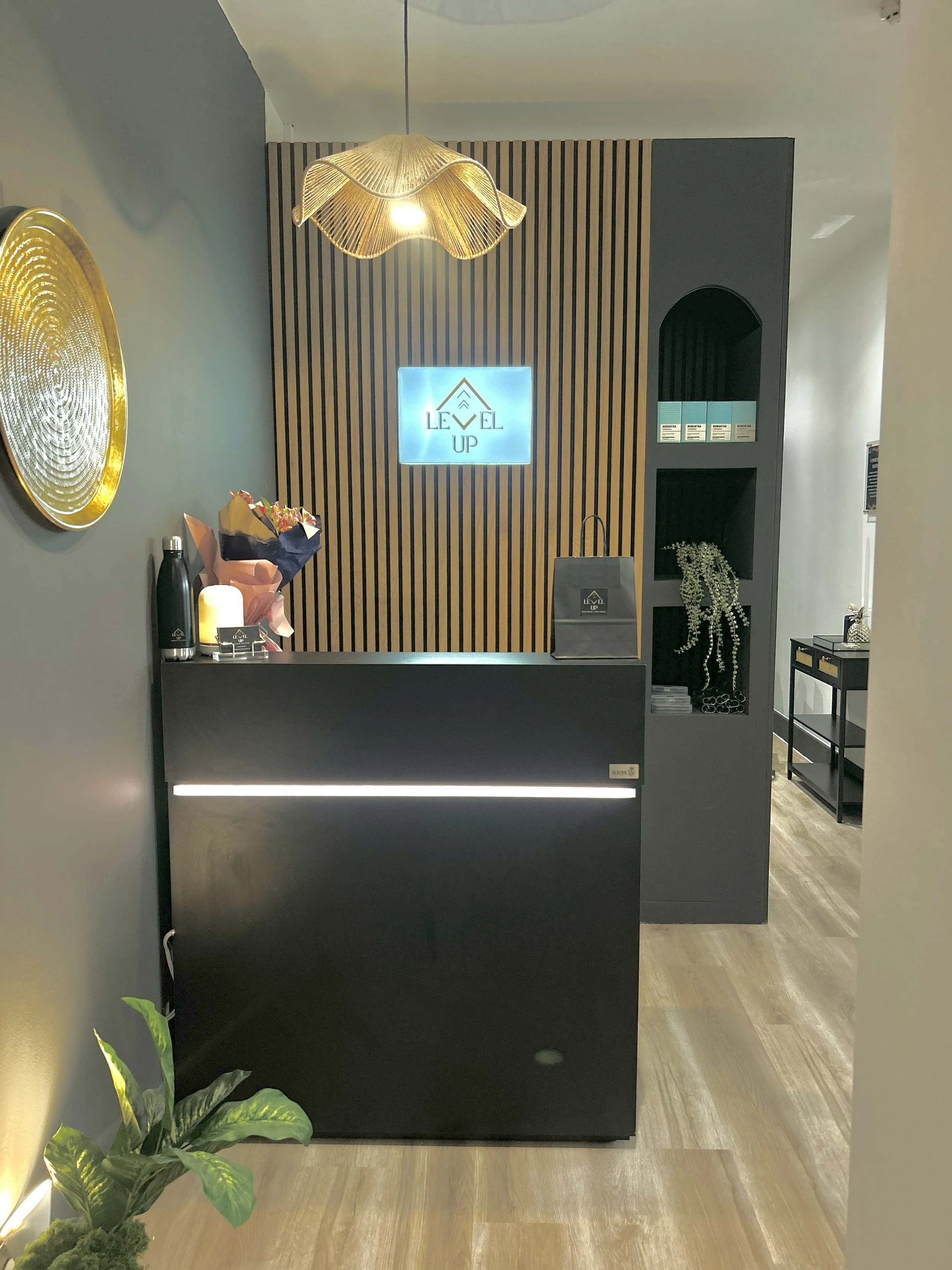 Modern reception desk with a black front panel, a floral bouquet, a black water bottle, and a lit candle on top, against a wall with a wooden slat accent and a sign that reads 'LEVEL UP.' There is a decorative plant in the lower left corner and a sma