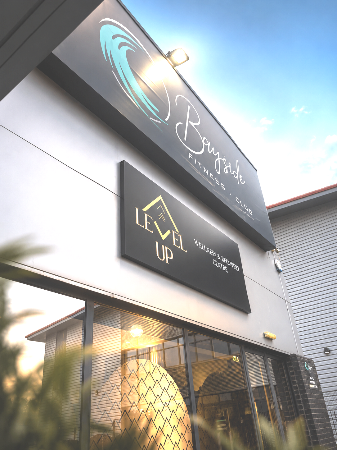 Exterior of a fitness club and wellness center with signs reading "Bayside Fitness Club" and "Level Up Wellness & Recovery Centre" during sunset.