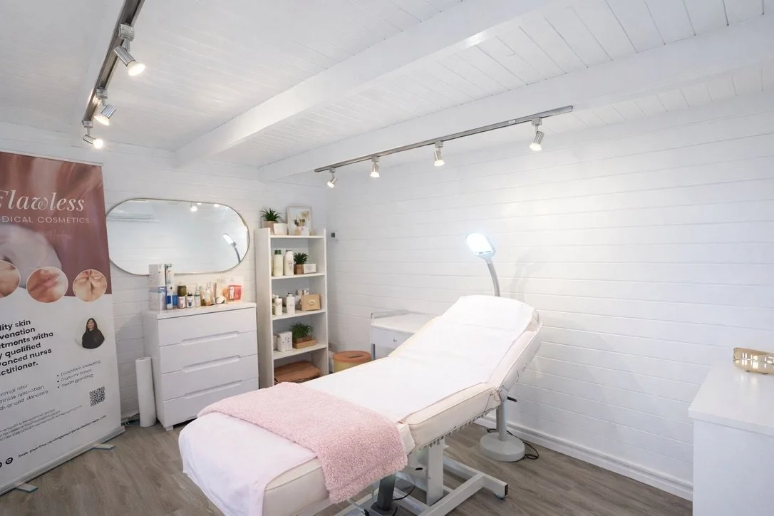 A clean, minimal aesthetic treatment room with a white treatment bed covered with a pink towel, surrounded by white furniture and shelves filled with skincare products, illuminated by overhead and adjustable lighting.