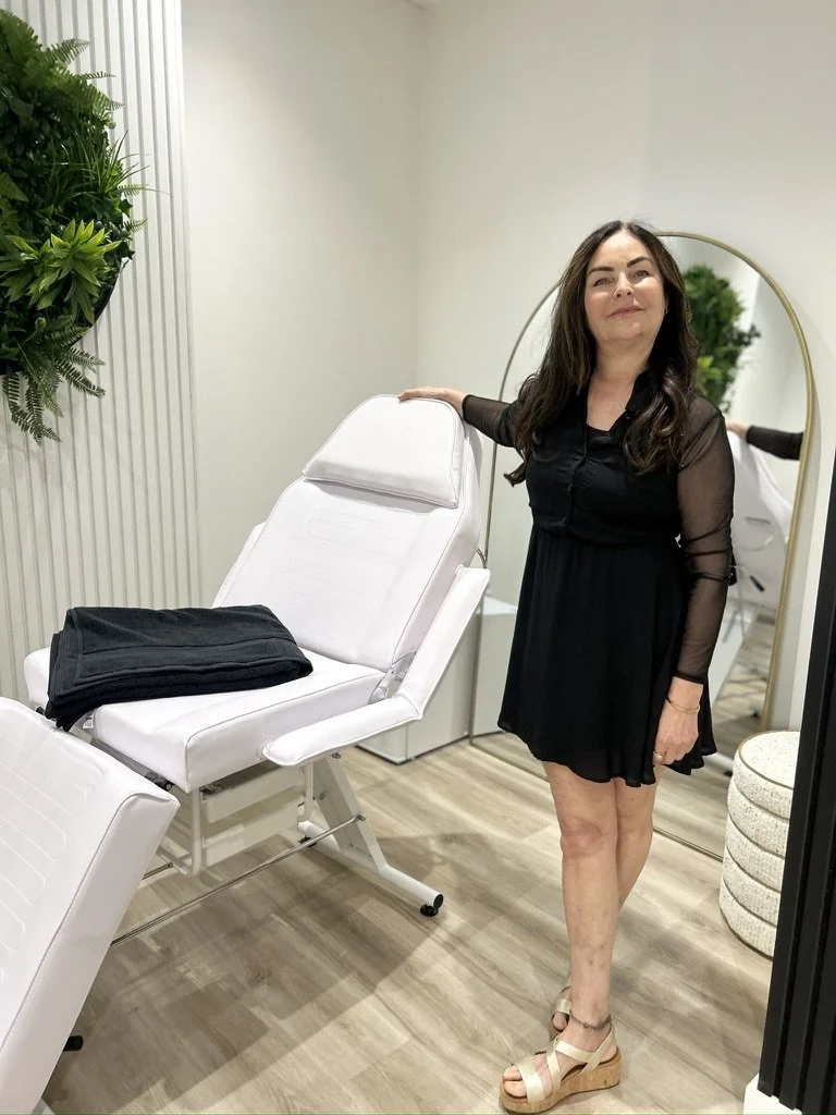 Woman in black dress and beige sandals standing next to a white medical or spa treatment chair in a modern treatment room with a full-length mirror and green plants.