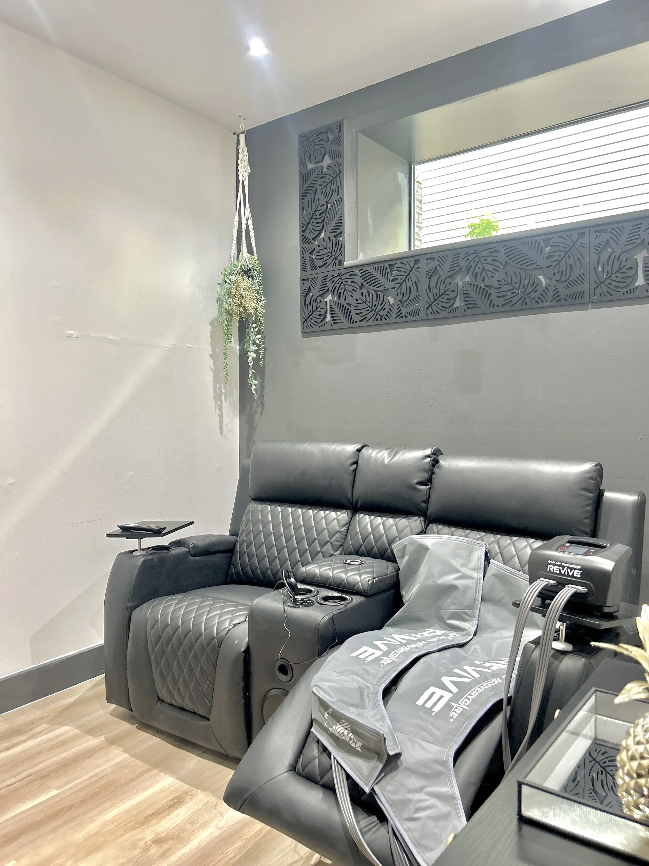 A living room features a black leather massage chair with quilted details, a small side table with a remote control, and a large window with decorative leaf-pattern acrylic panels. A hanging plant decorates the corner, and there's a Bobath branding b