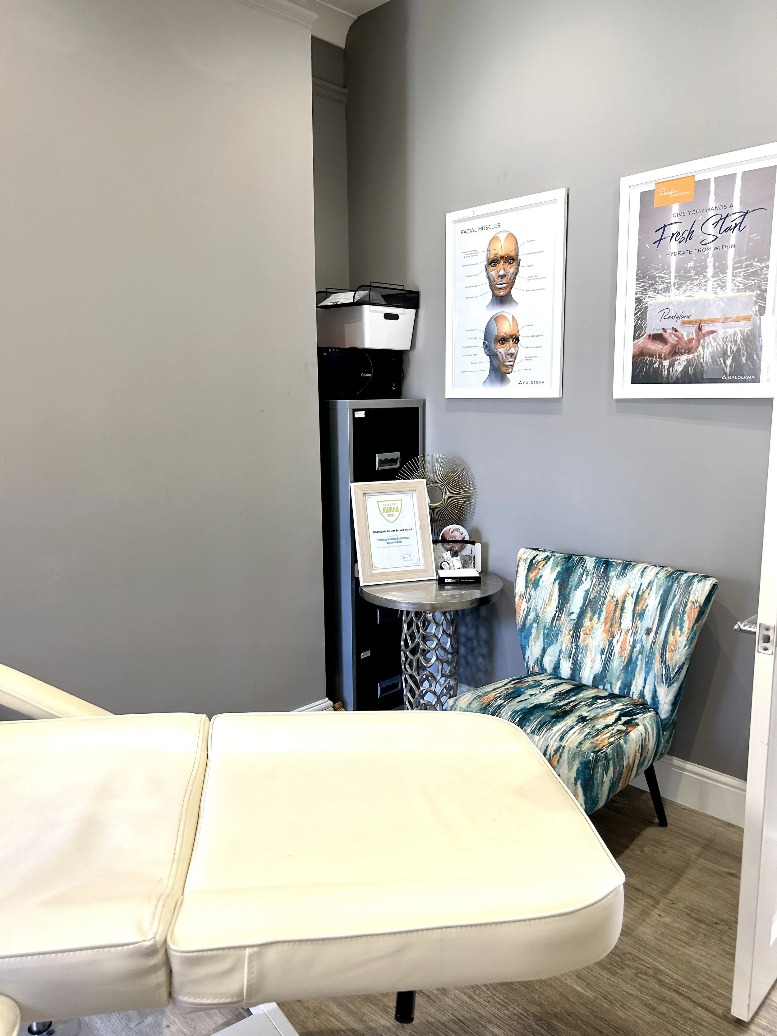 A room with a pale yellow upholstered chair, a decorated armchair with a multicolored abstract pattern, a gray wall, and framed posters on the wall featuring facial muscles and skincare advertisements. There is a small side table with awards, certifi