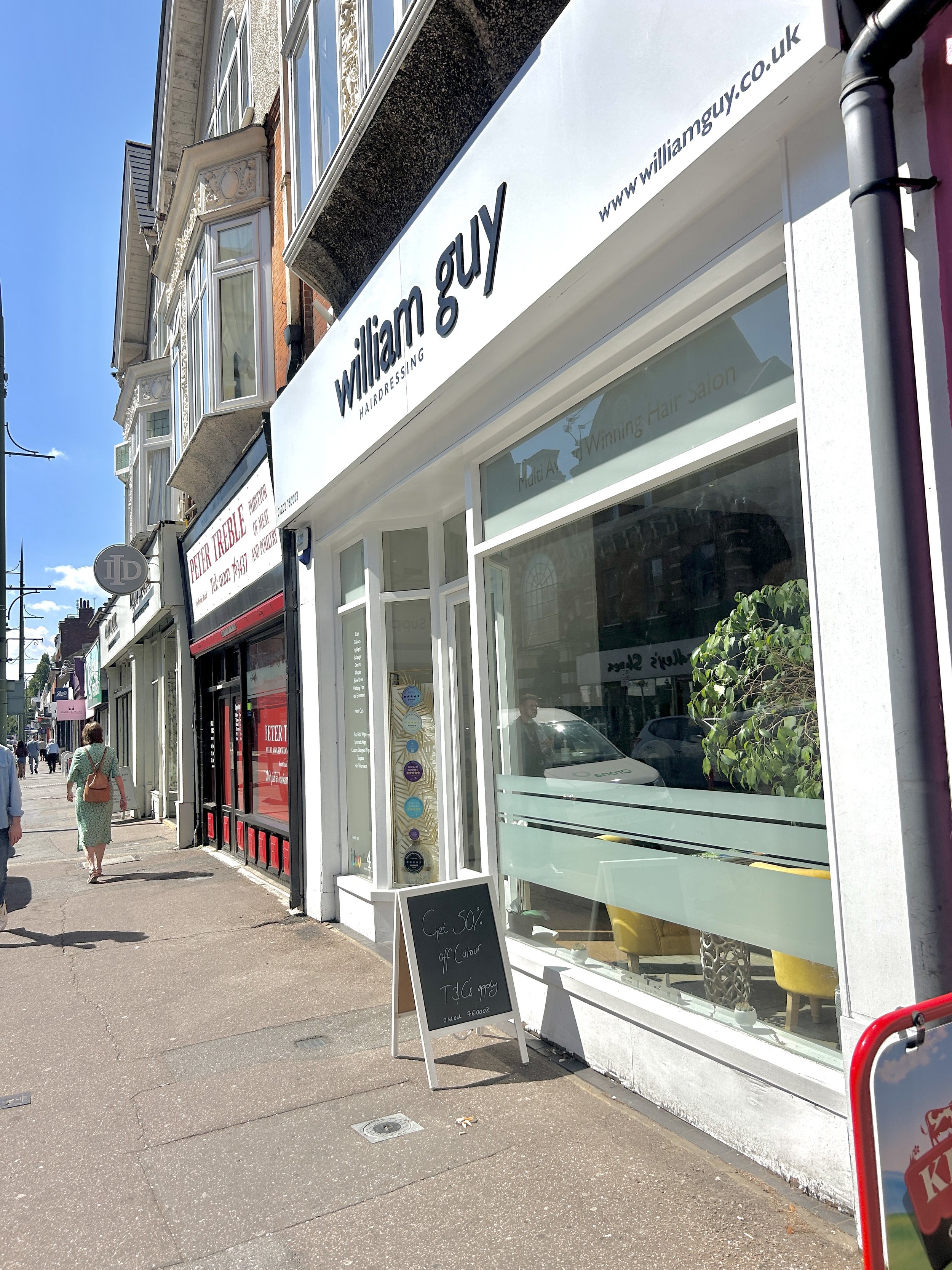 View of a storefront with a sign that reads 'William Guy Hairdressing' and a black sandwich board displaying a 50% off promotion, on a busy city street with pedestrians and buildings.