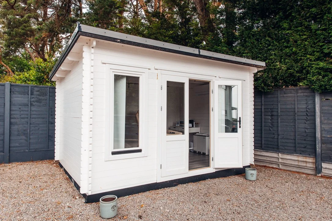 Small white garden shed with large glass doors and windows, situated on gravel ground, enclosed by a dark wooden fence with trees in the background.