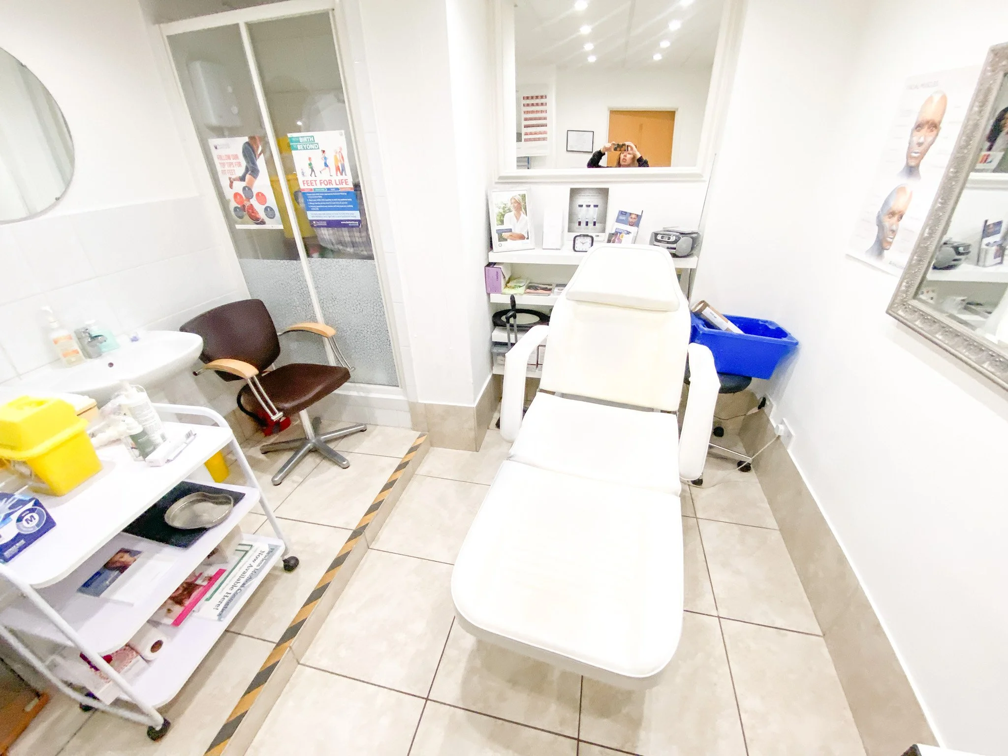 A small medical examination room with white walls and beige tiled floors. There is a white examination table, a white chair, and a brown chair. Medical supplies and informational materials are on a white cart and shelves. A mirror on the wall reflect