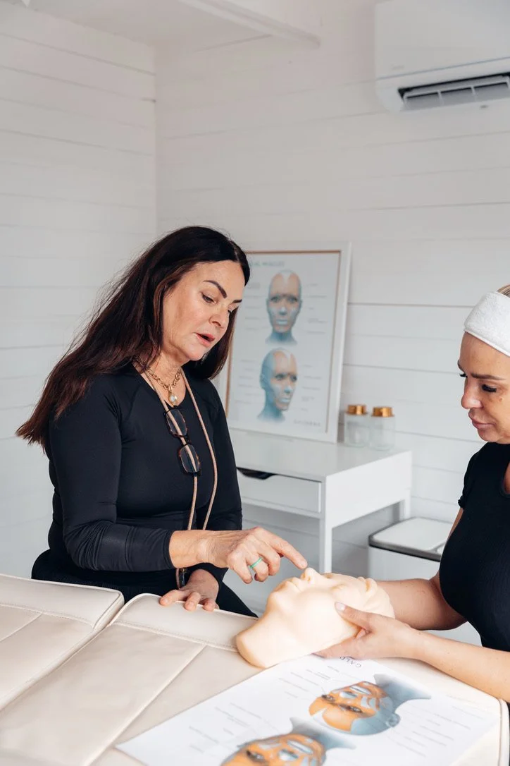 Two women in a clinical setting discussing a facial mannequin, with one woman pointing at the mannequin's nose. In the background, there is a medical chart and a white cabinet with small containers.