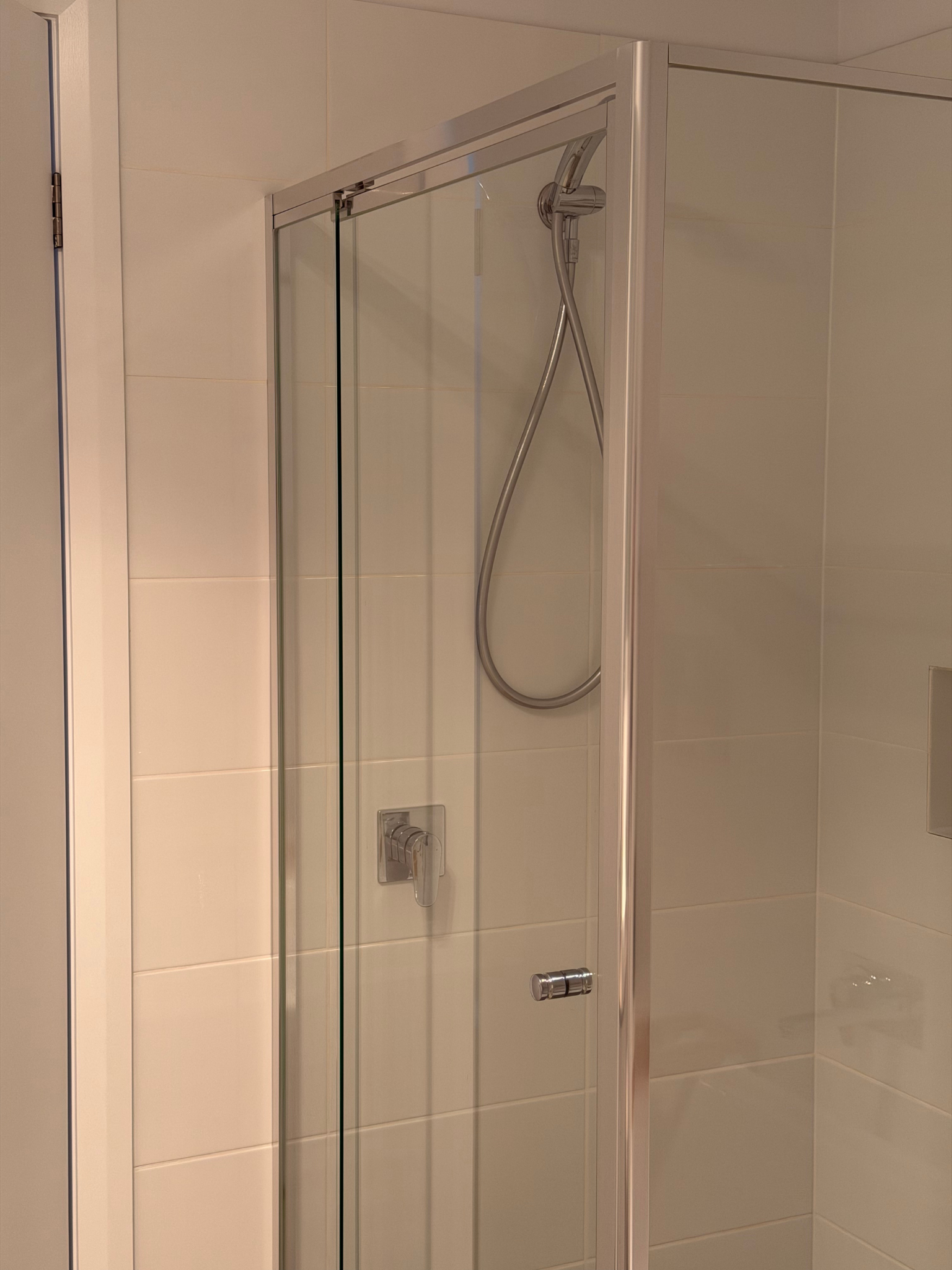 A corner shower with white tiled walls, a glass door, and a chrome showerhead with a flexible hose inside the shower enclosure.