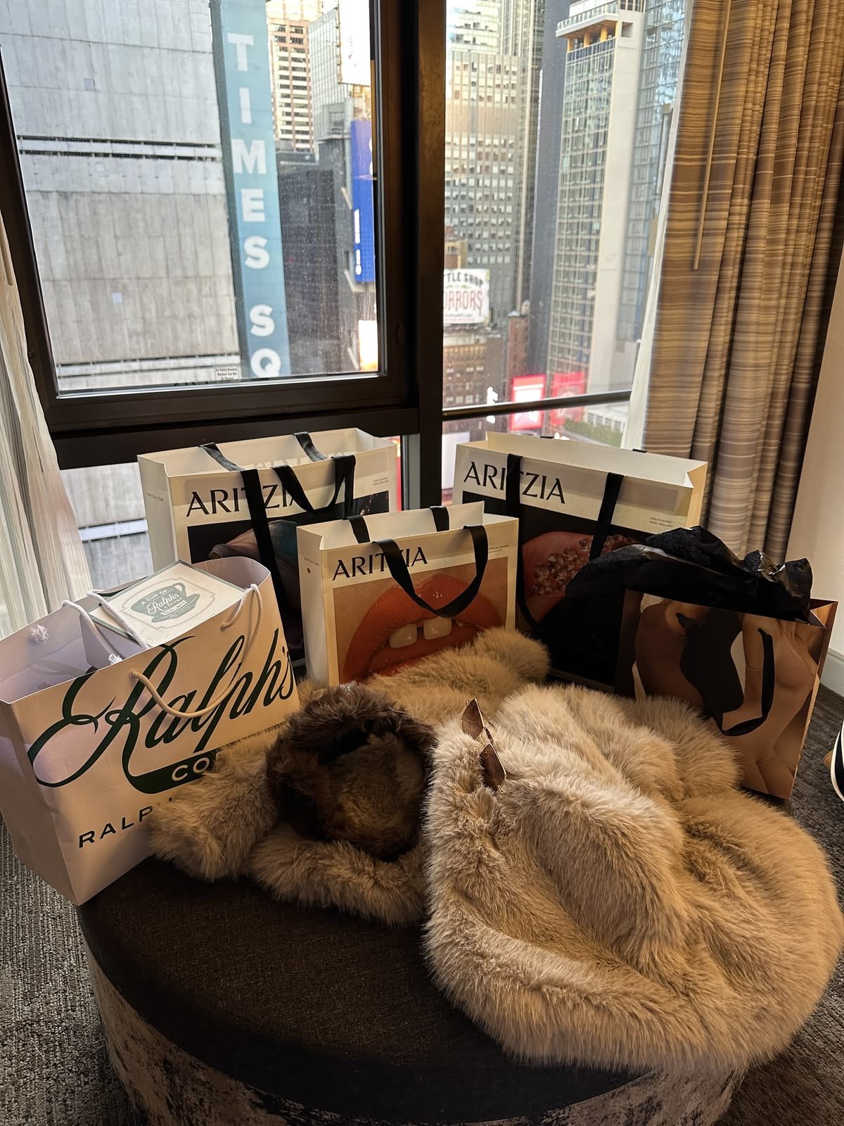 A person with long hair is lying on a fur blanket on a round ottoman in front of a window with a city view. Several shopping bags from Ralph's, AriZia, and other stores are placed on the ottoman.