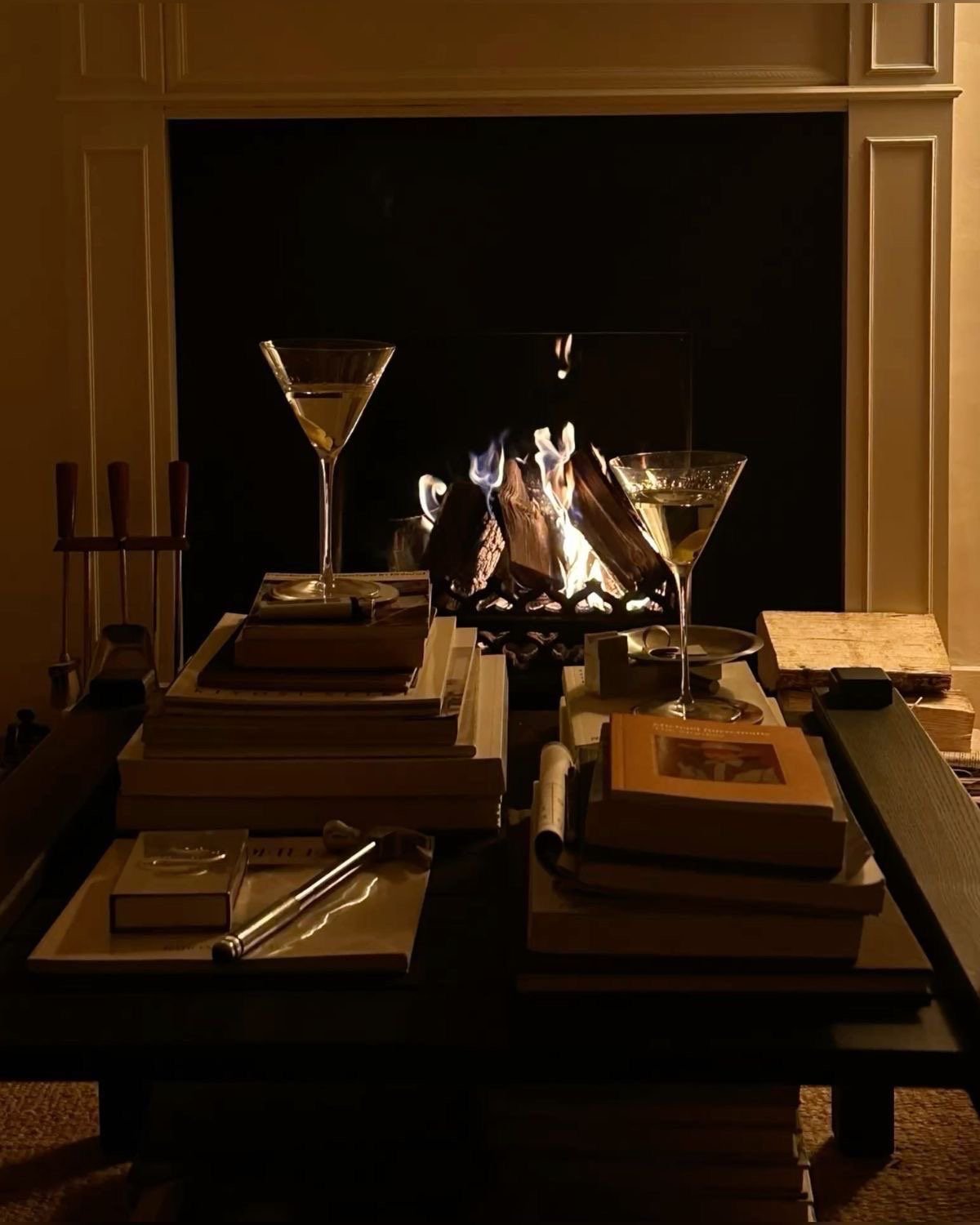 Fireplace in a room with a lit fire and logs, surrounded by books, notebooks, and two martini glasses on a table.
