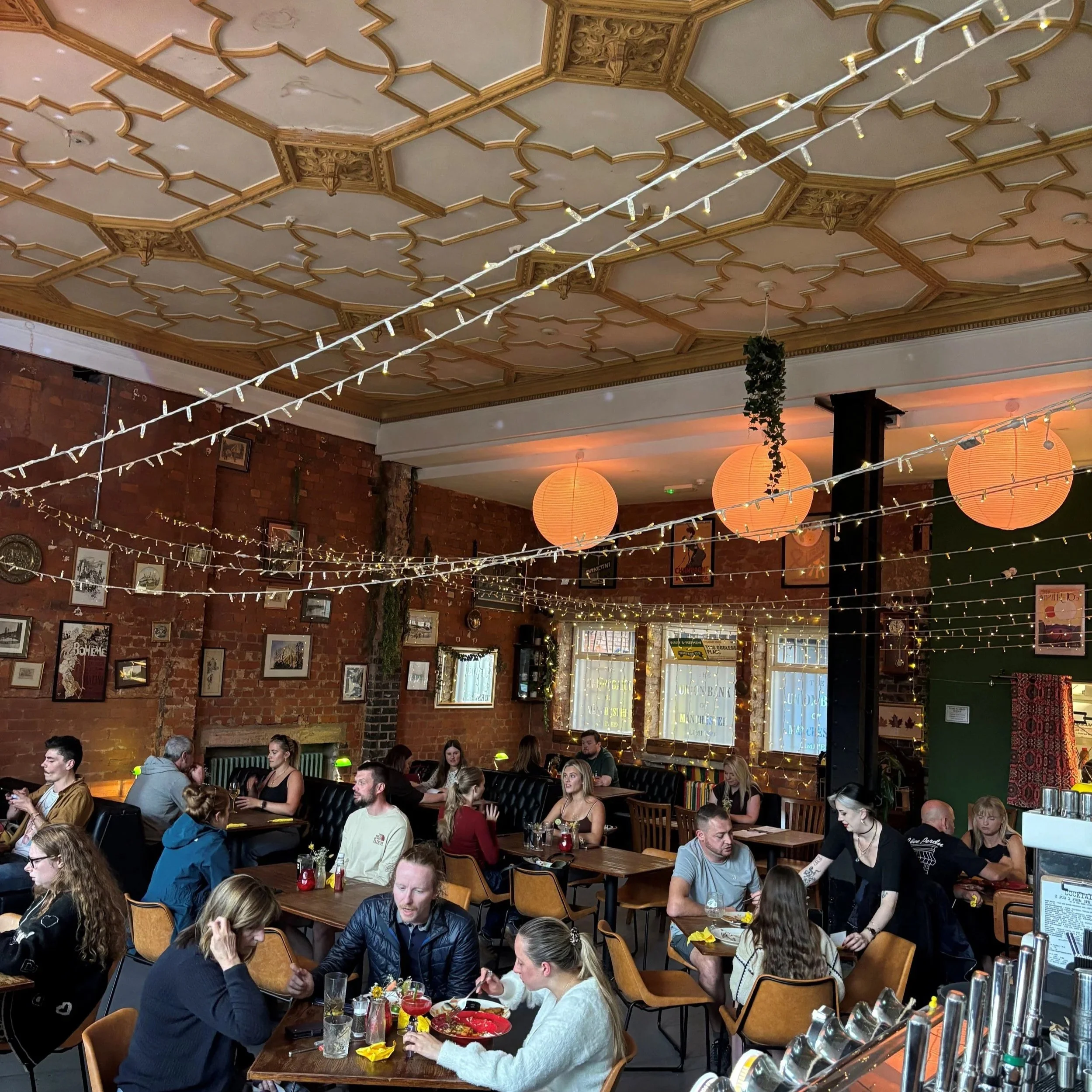 People dining at tables in a restaurant with brick walls, decorated with framed pictures, and a wooden ceiling with ornate molding. String lights and hanging paper lanterns create a warm atmosphere.