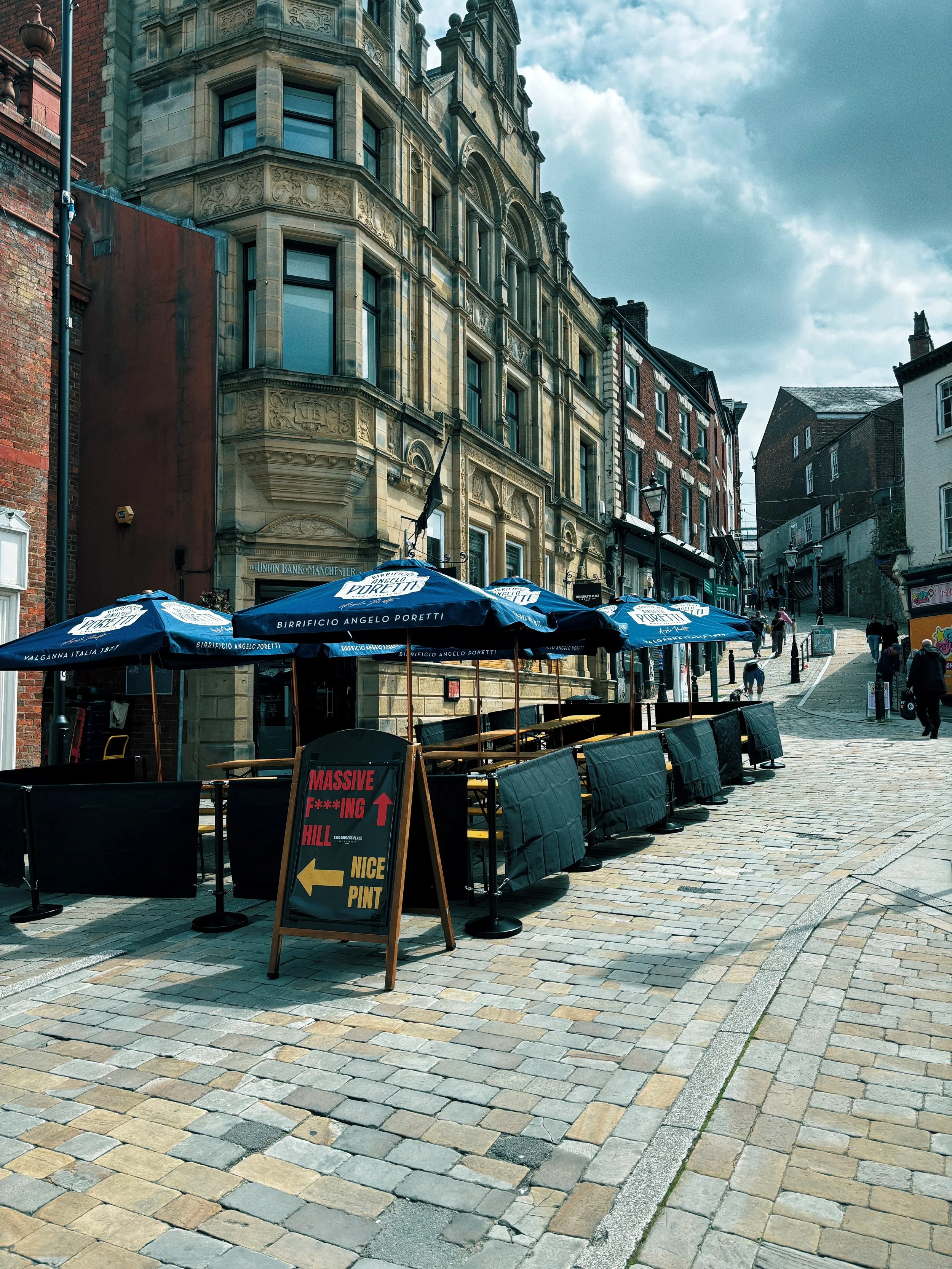 A street scene with historic multi-story buildings, outdoor umbrellas for a patio, and a chalkboard sign with directional information, cobblestone pavement, and pedestrians walking up a hill.