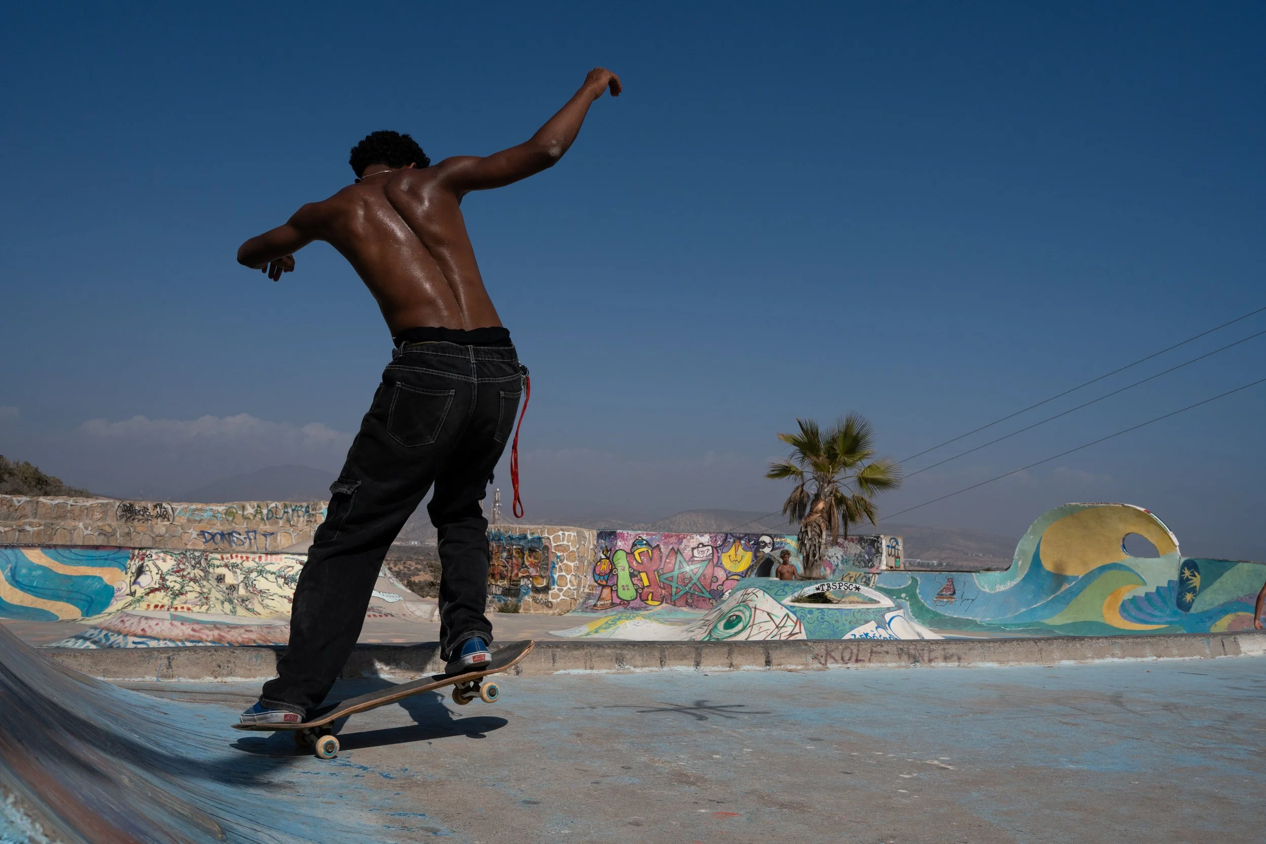 A shirtless person skateboarding at a graffiti-covered skate park under a clear blue sky.