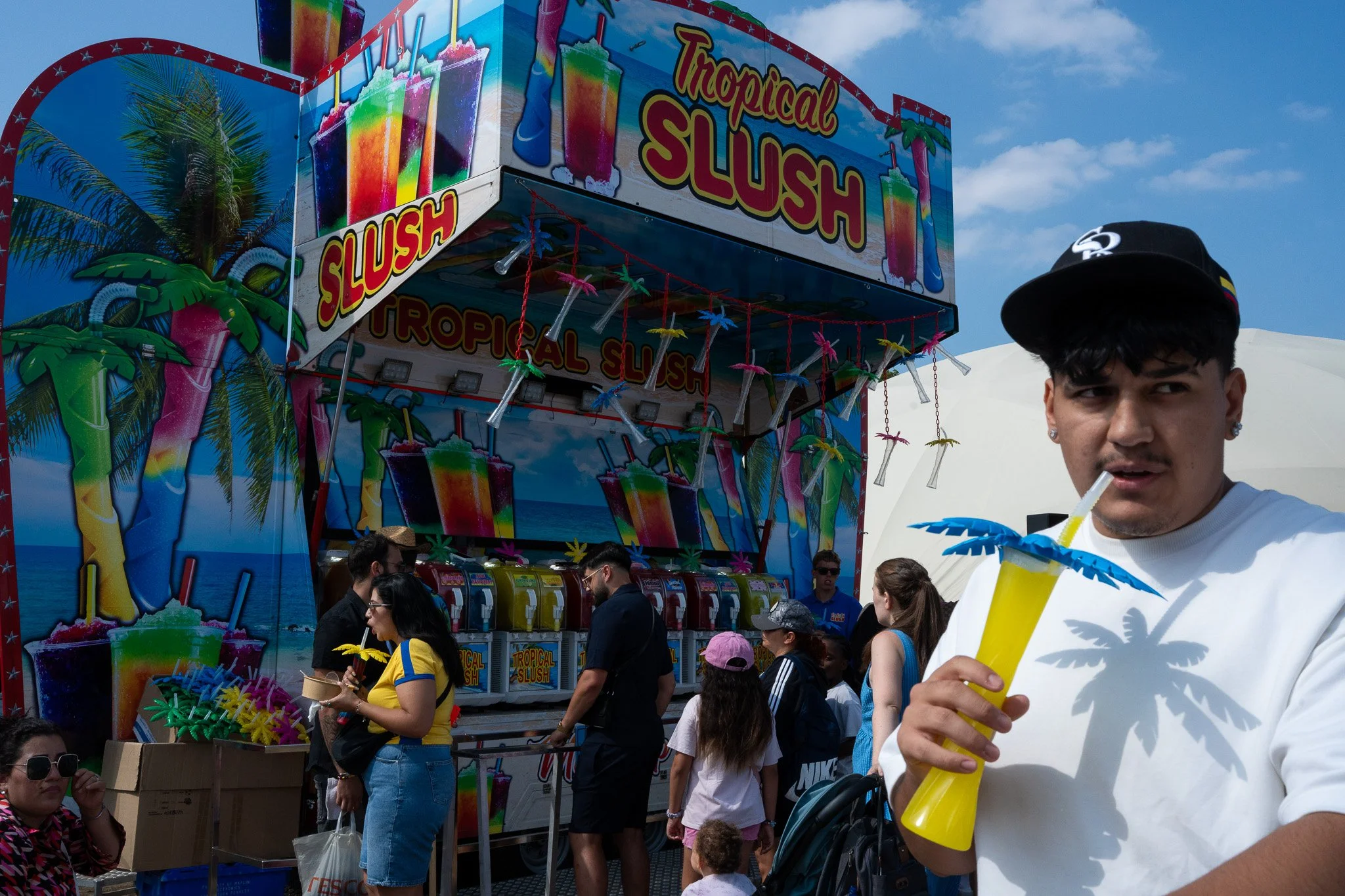 A crowded outdoor event with a colorful vendor booth labeled 'Tropical Slush' featuring images of rainbow-colored slush drinks and palm trees. People are gathered around, some holding yellow drinks with umbrellas, and a man in a black cap and white s