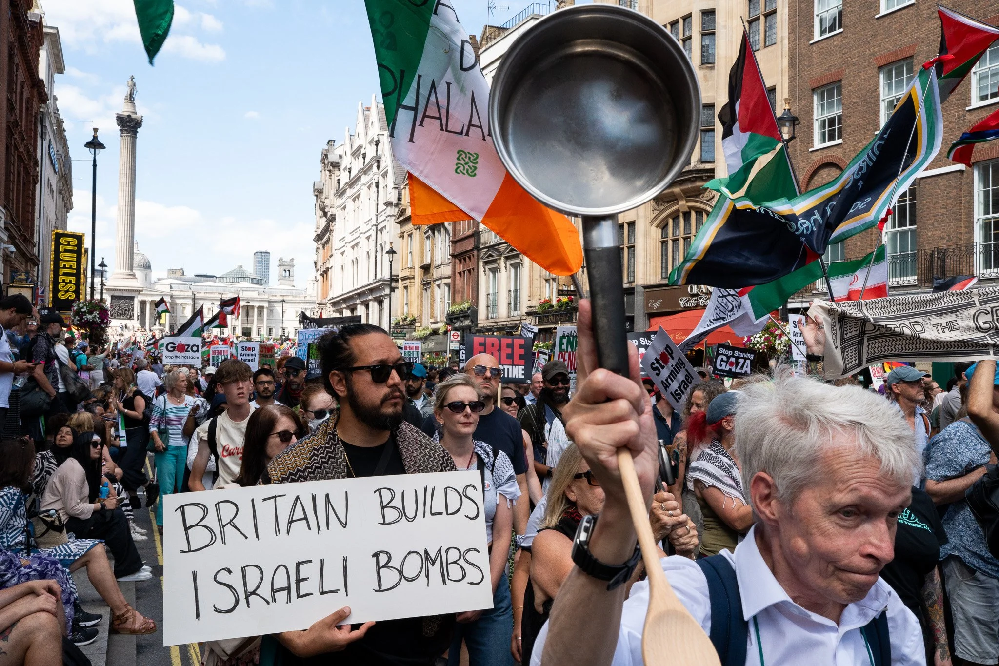 Crowd of protesters in a London street holding signs and flags, including a sign that reads 'Britain builds Israeli bombs'. A person holds a metal pot above the crowd.