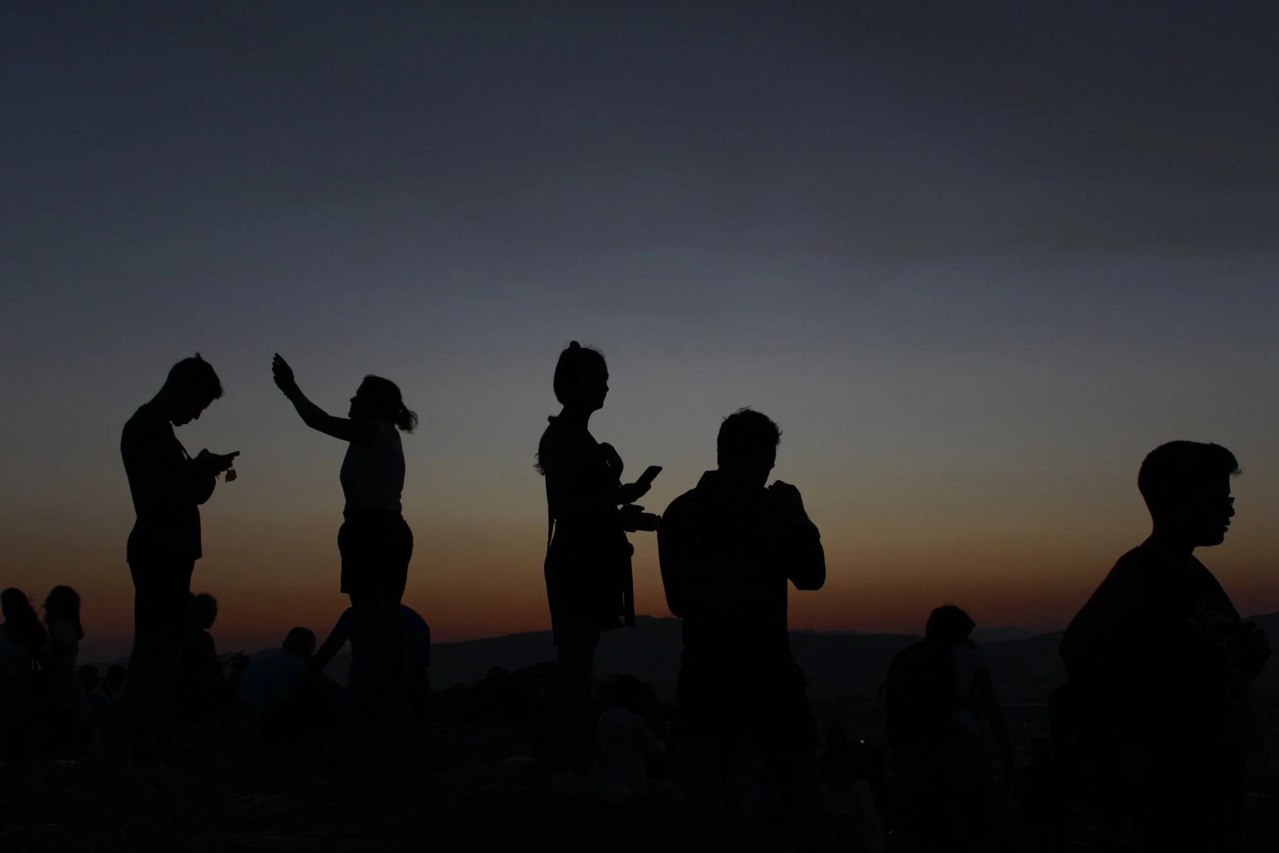 Silhouettes of people standing and sitting outdoors during sunset, with some using mobile devices, against a gradient sky