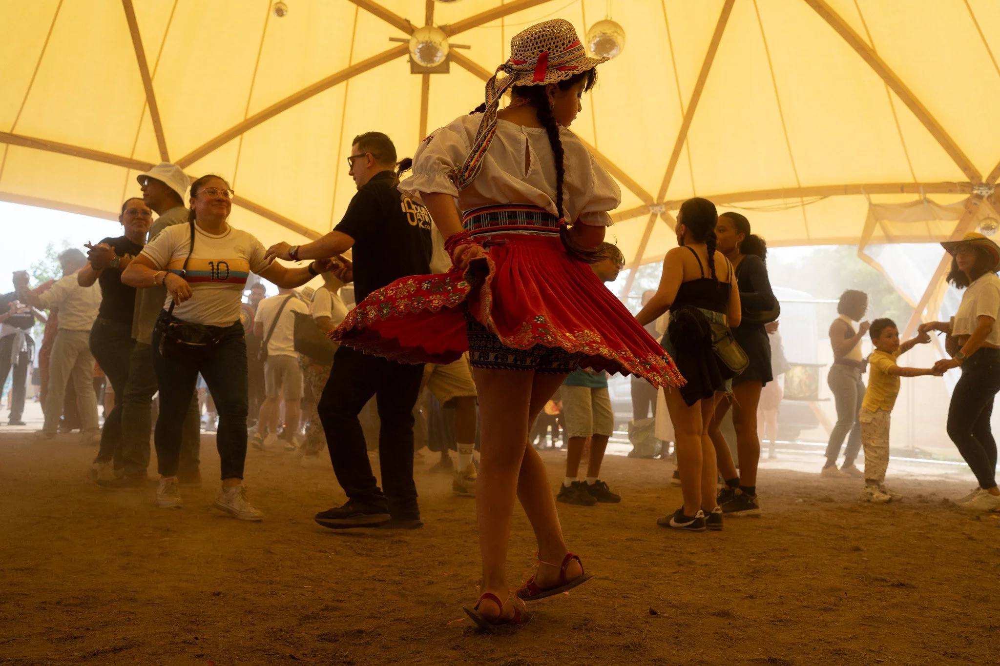 A girl in a traditional folk costume dancing in a circle at an indoor cultural event, with other people dancing and socializing under a large yellow tent.