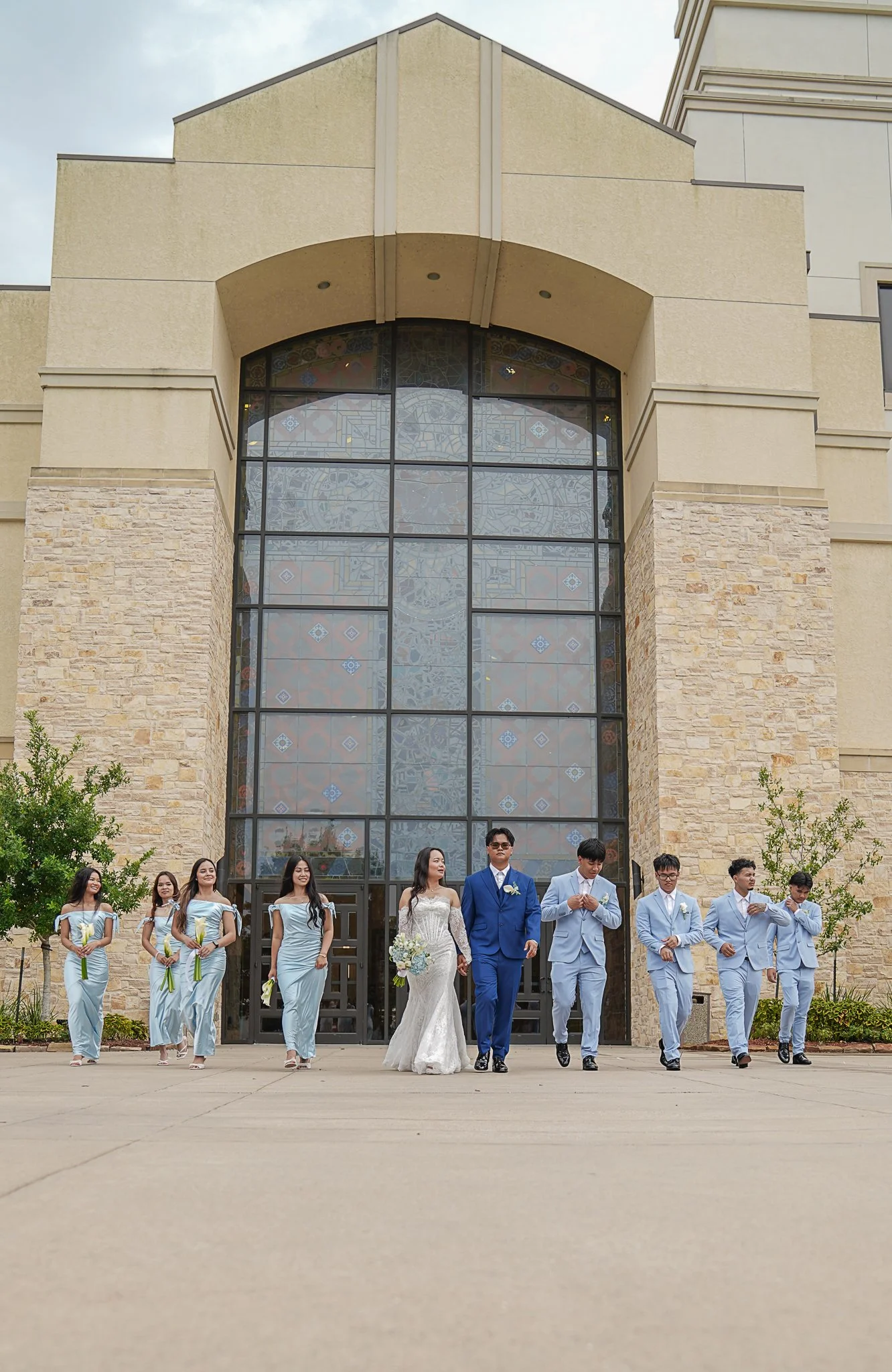 A bride and groom walking out of a church with wedding party members and bridesmaids and groomsmen beside them.