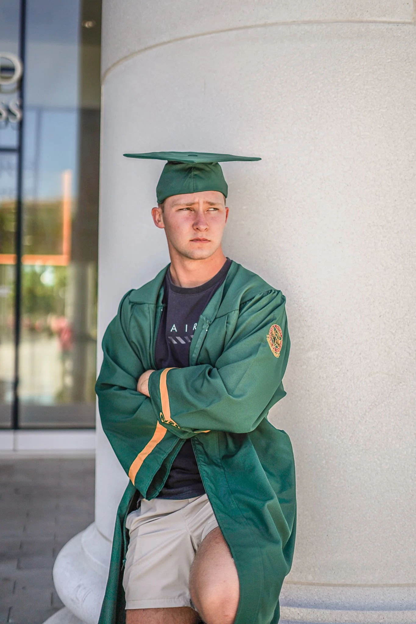A man wearing a green graduation cap and gown, with crossed arms, standing outdoors against a light-colored wall, looking to the side.