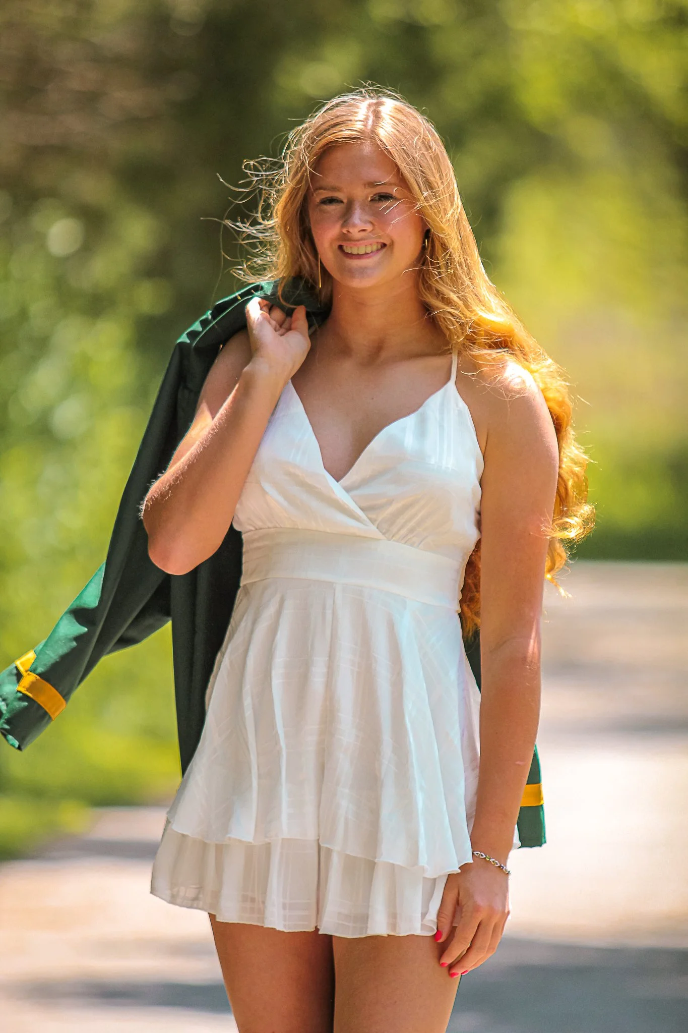 A woman with long red hair wearing a white dress with a pleated skirt, holding a black jacket over her shoulder, standing outdoors on a sunny day.
