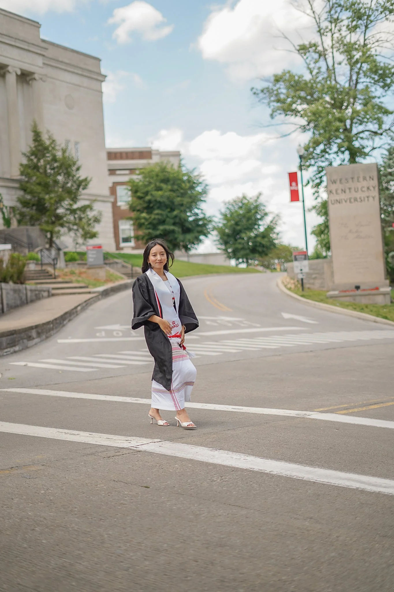 A woman in formal attire, wearing a graduation gown and sash, walking across a street near Western Kentucky University campus entrance, with trees and campus buildings in the background on a partly cloudy day.