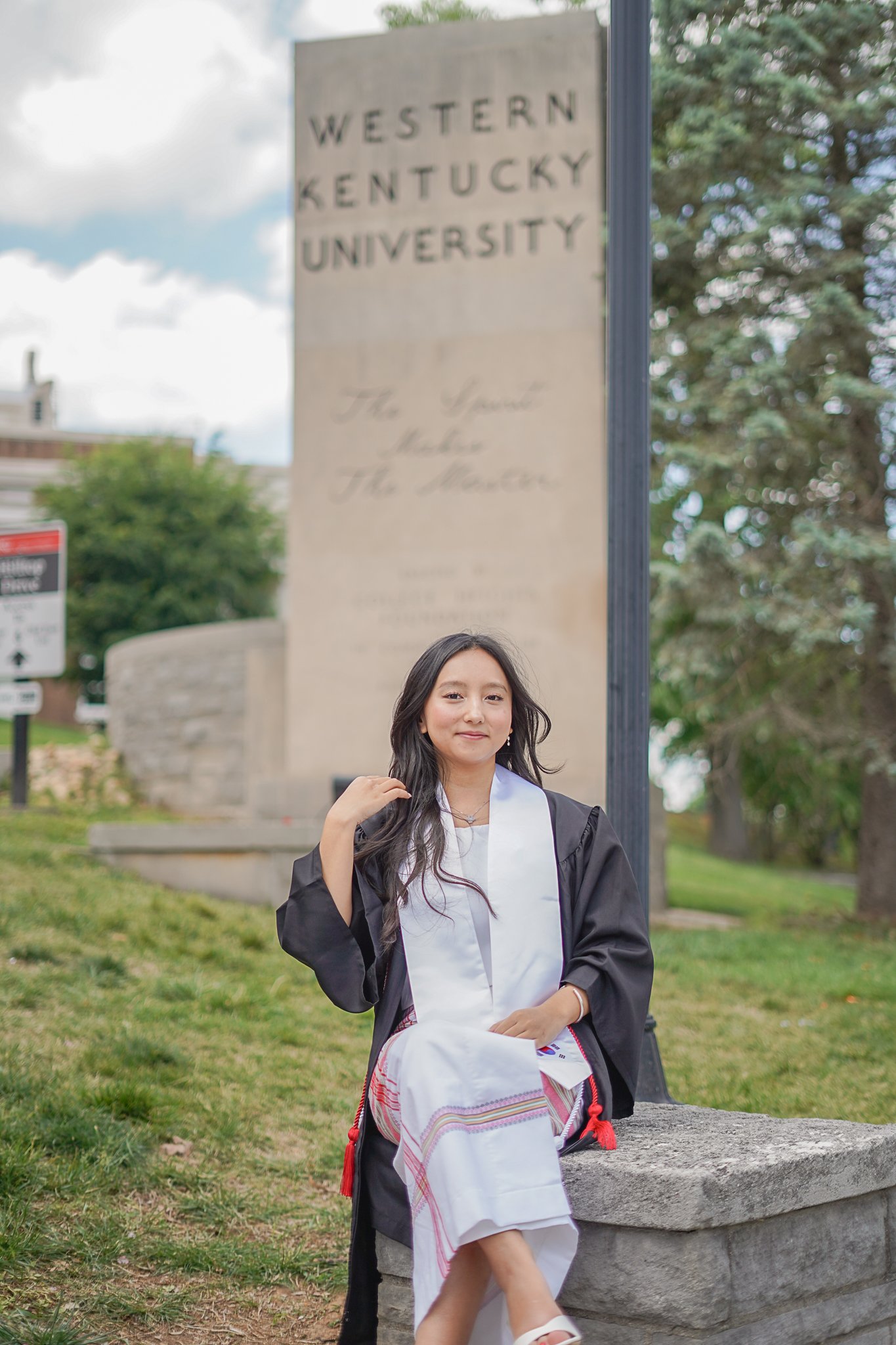 A young woman in graduation attire, including a black cap and gown, sitting on a stone bench outdoors near a large sign that reads 'Western Kentucky University'. She has long dark hair, is smiling slightly, and has a white sash or stole over her gown