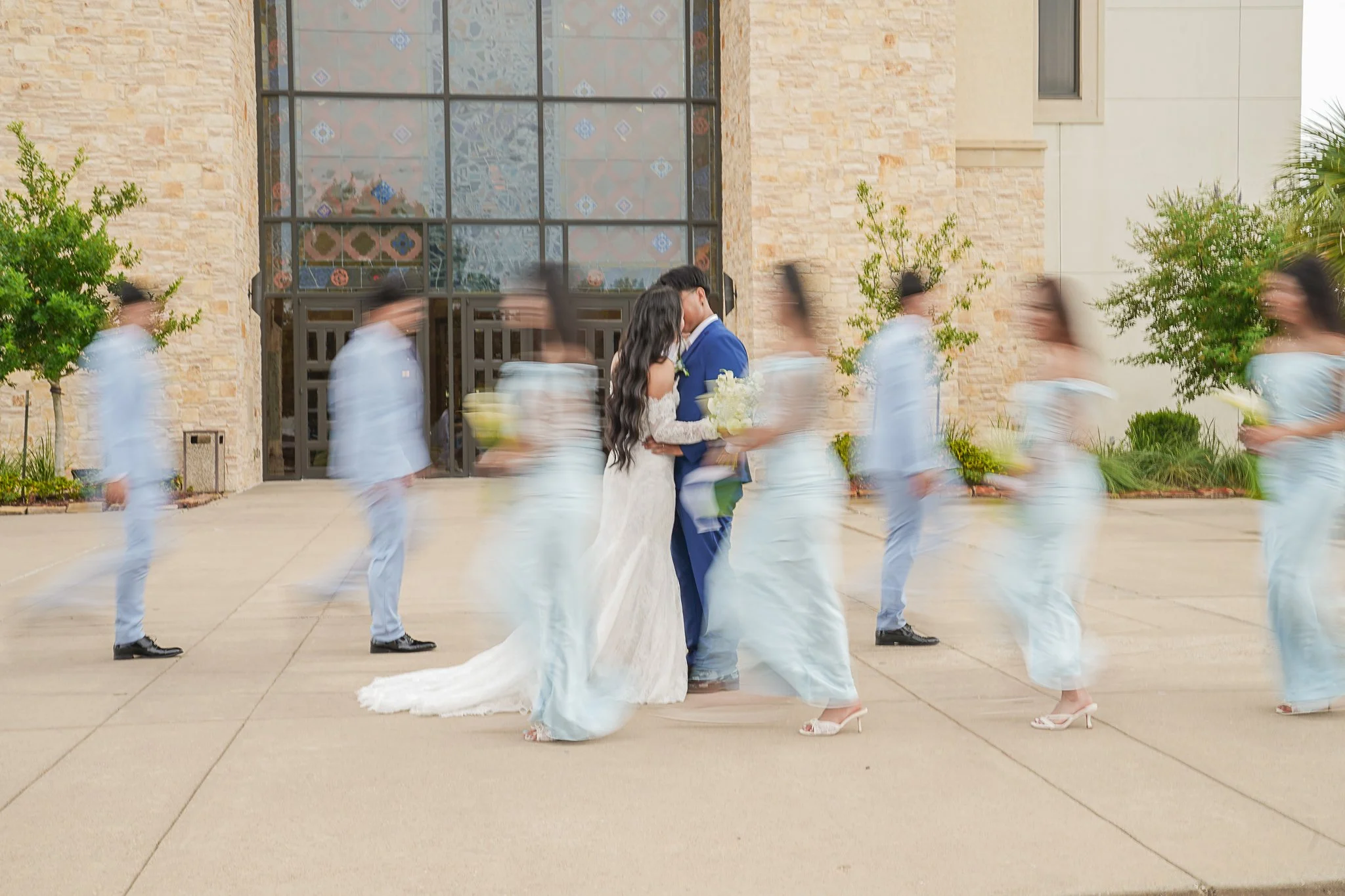 A bride and groom touching foreheads at their wedding with dancers dressed in white around them, in front of a stone building with large stained glass windows.