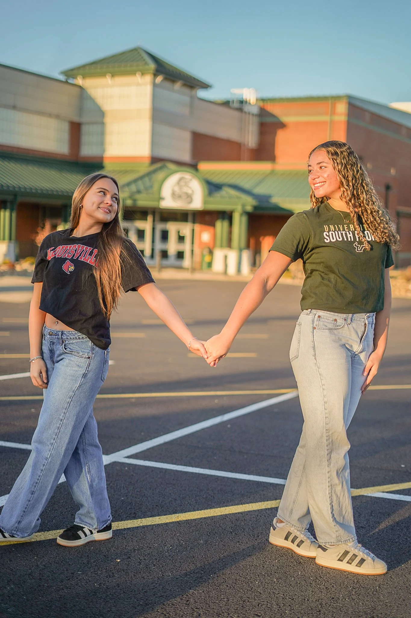 Two young women holding hands and smiling in a parking lot outside a building.