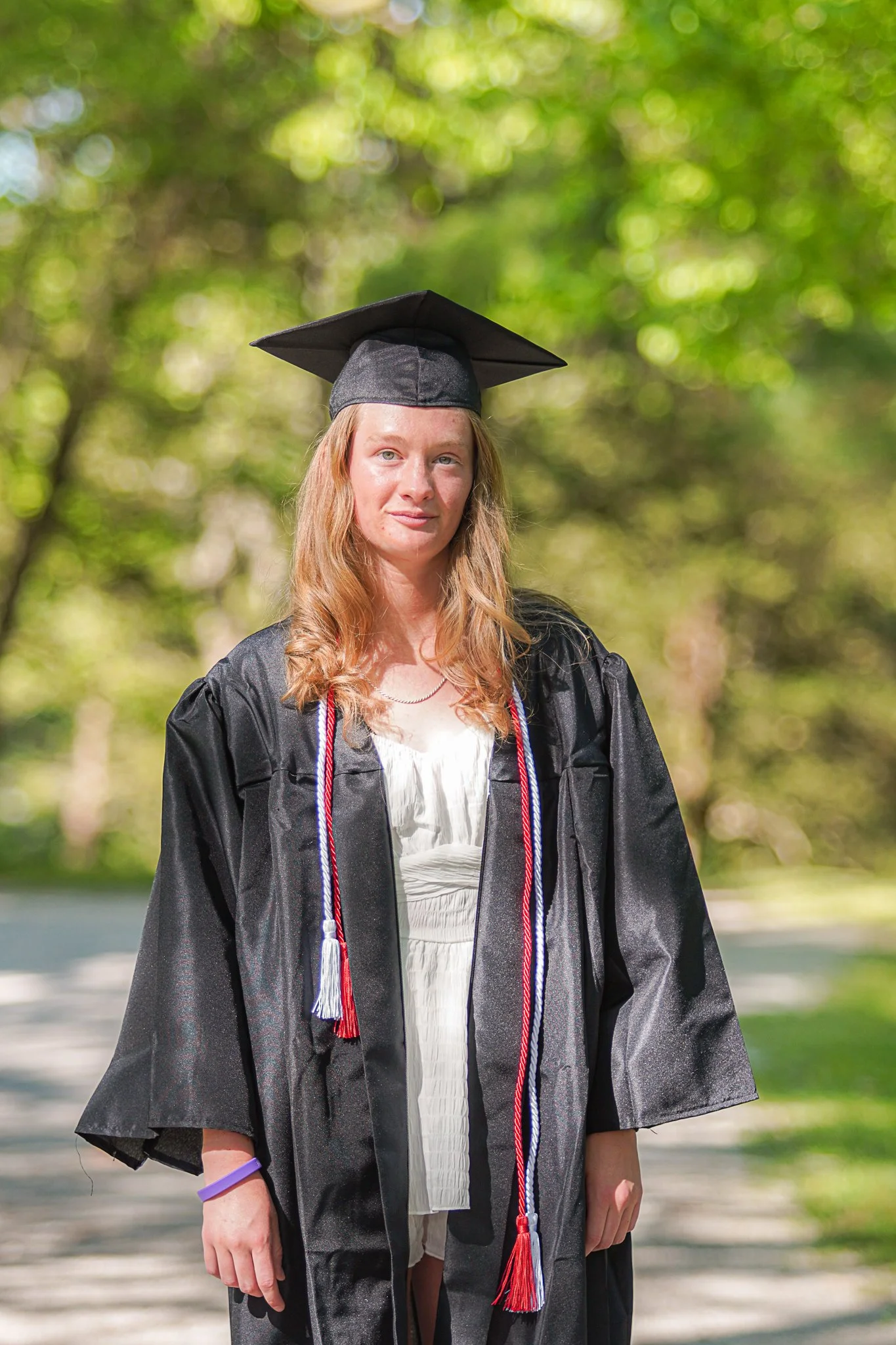 A young woman in a black graduation gown and cap, with a red, white, and blue cord, standing outdoors in front of green trees.