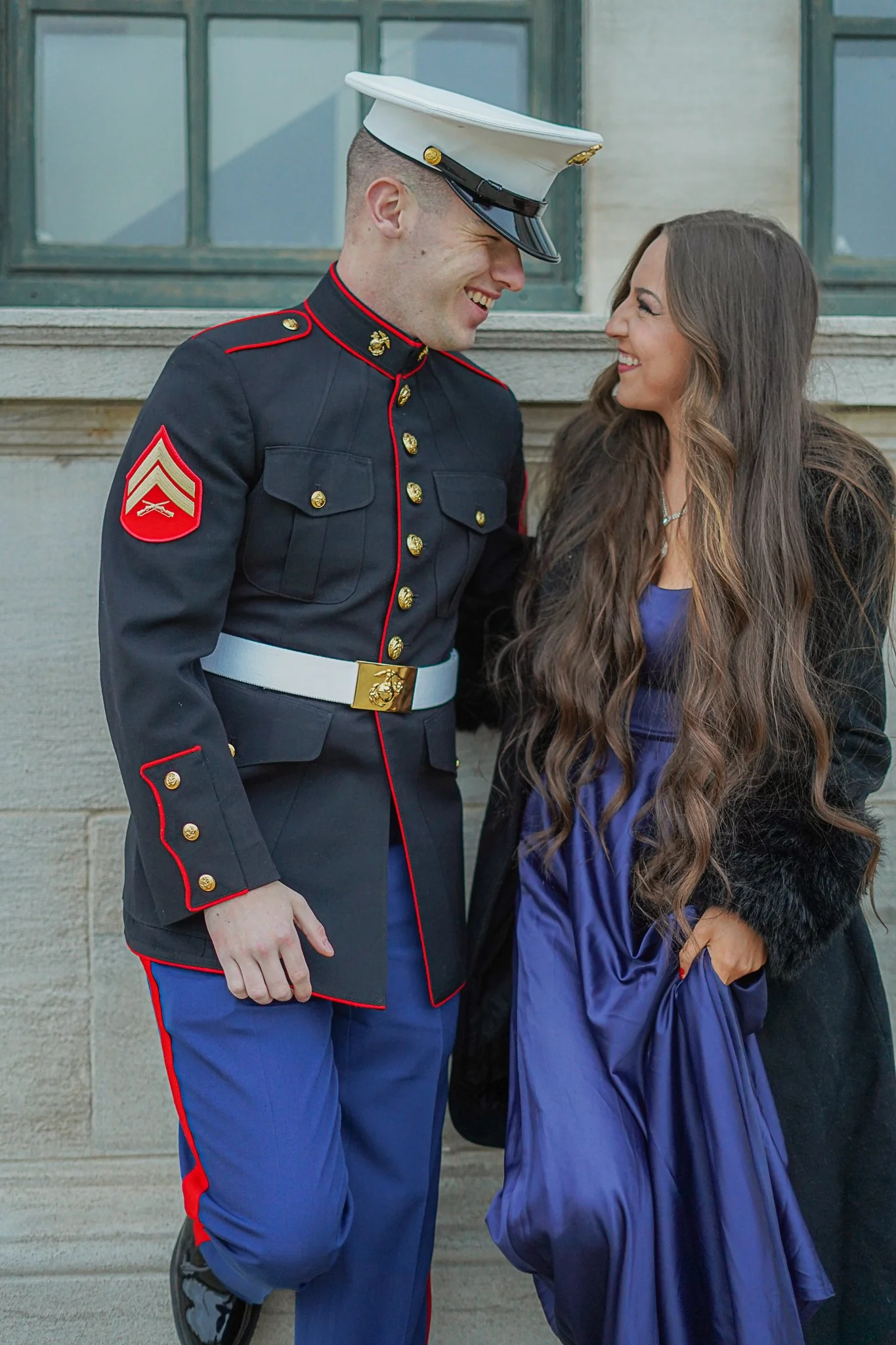 A young man in a United States Marine Corps dress uniform smiling and leaning toward a woman, who is also smiling and looking at him. They are standing outdoors in front of a building with large windows.