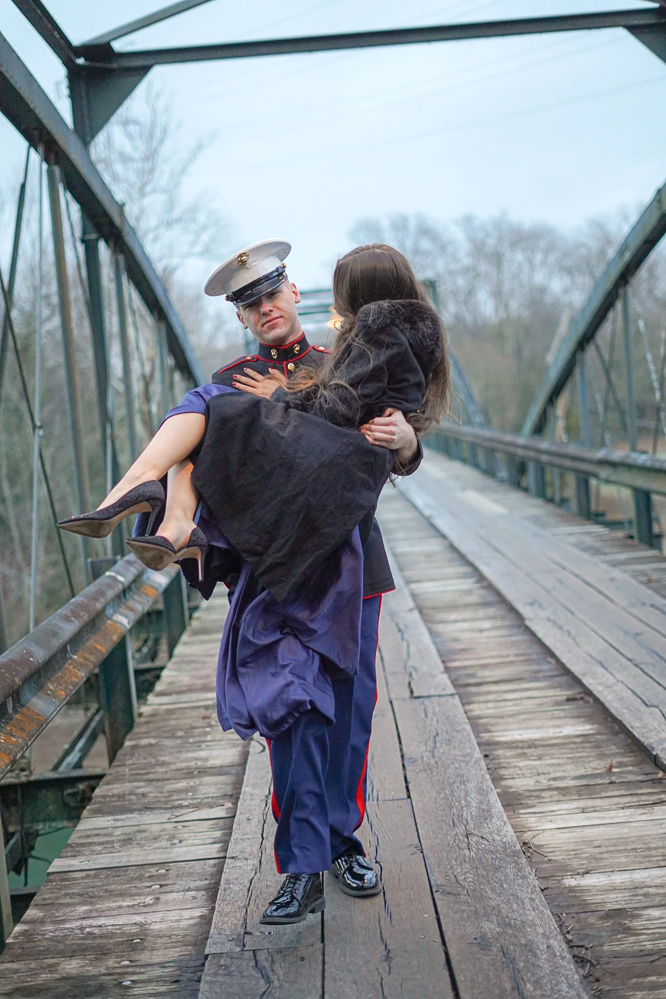 A man in a military uniform holding a woman in a black coat, on a wooden bridge with a metal railing, trees in the background, overcast sky.