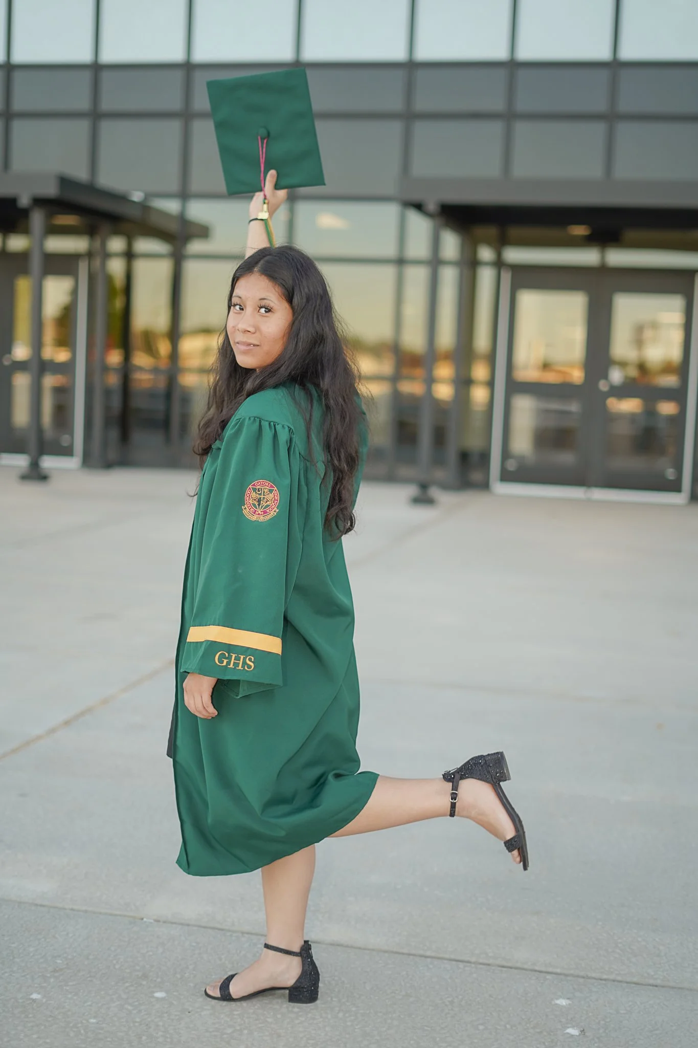 A young woman wearing a green graduation gown is outside in front of a modern glass building. She is holding her graduation cap high in the air with her right hand and is balancing on her left leg with her right leg bent at the knee, wearing black hi