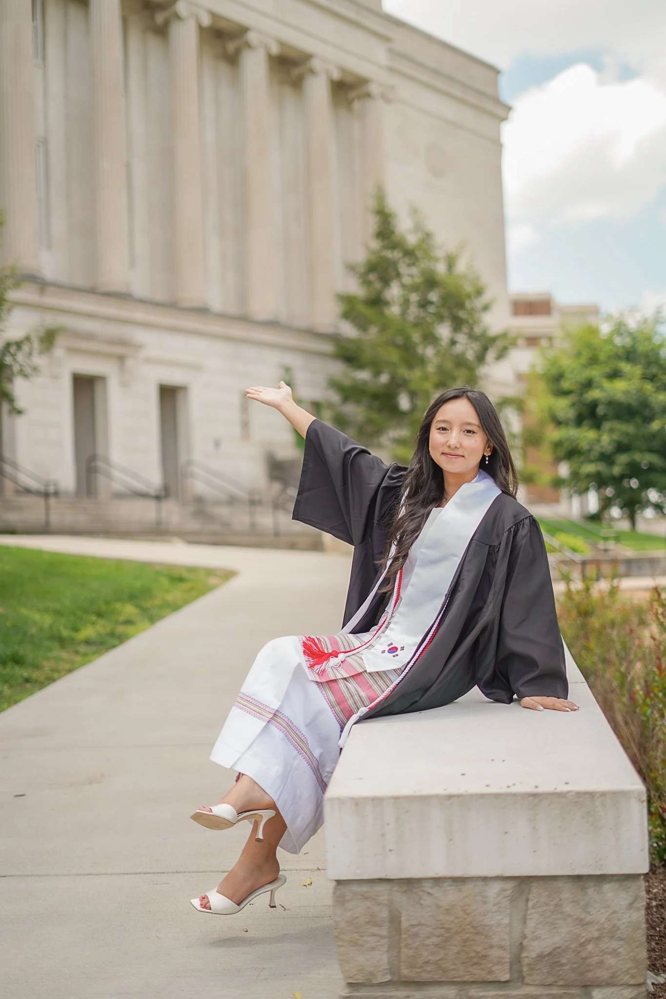 Young woman in a graduation gown sitting on a concrete ledge with her arm extended, outside a large classical building with columns, trees, and a blue sky with clouds.