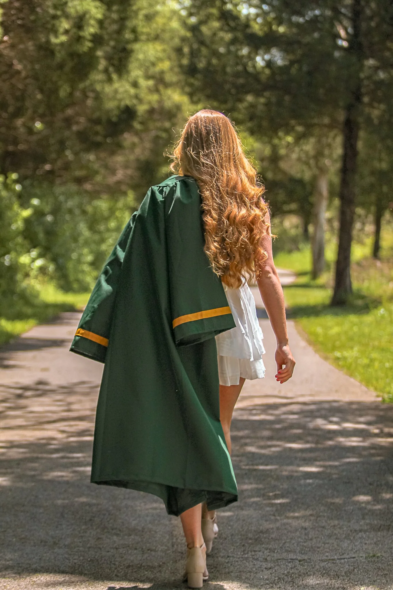 A woman walking on a sunny path in a park, dressed in a white dress and beige platform heels, with a green graduation gown draped over her shoulder and loose curly hair.