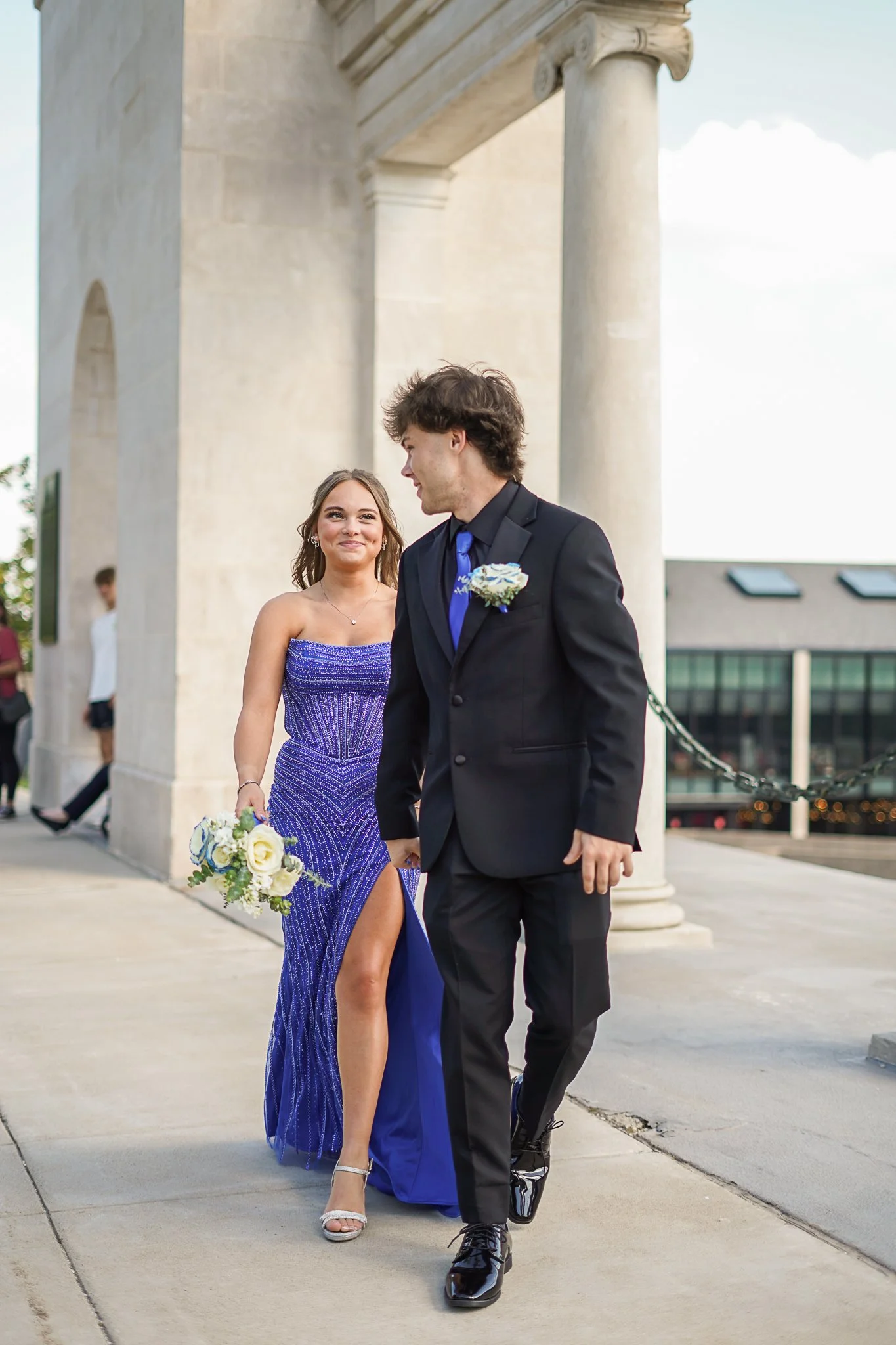 A young woman in a strapless blue dress holding a bouquet of white and blue flowers, walking beside a young man in a black suit with a blue tie and boutonnière, in front of a stone archway.