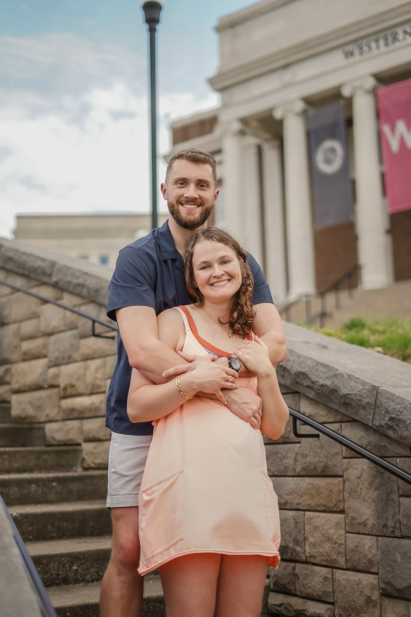 A happy young couple, the man with a beard wearing a navy blue shirt and the woman with curly hair in a peach dress, standing on outdoor stone stairs in front of a building with columns. The woman holds her belly, indicating pregnancy, and they are s