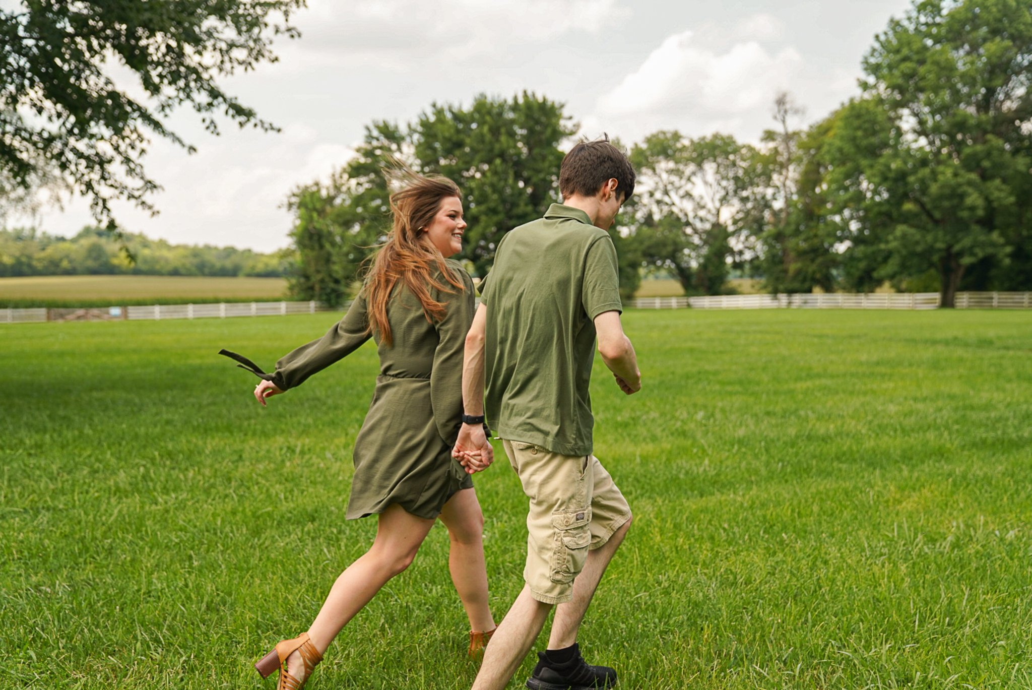 A young couple holding hands and walking together in a grassy field on a cloudy day, smiling and enjoying each other's company.