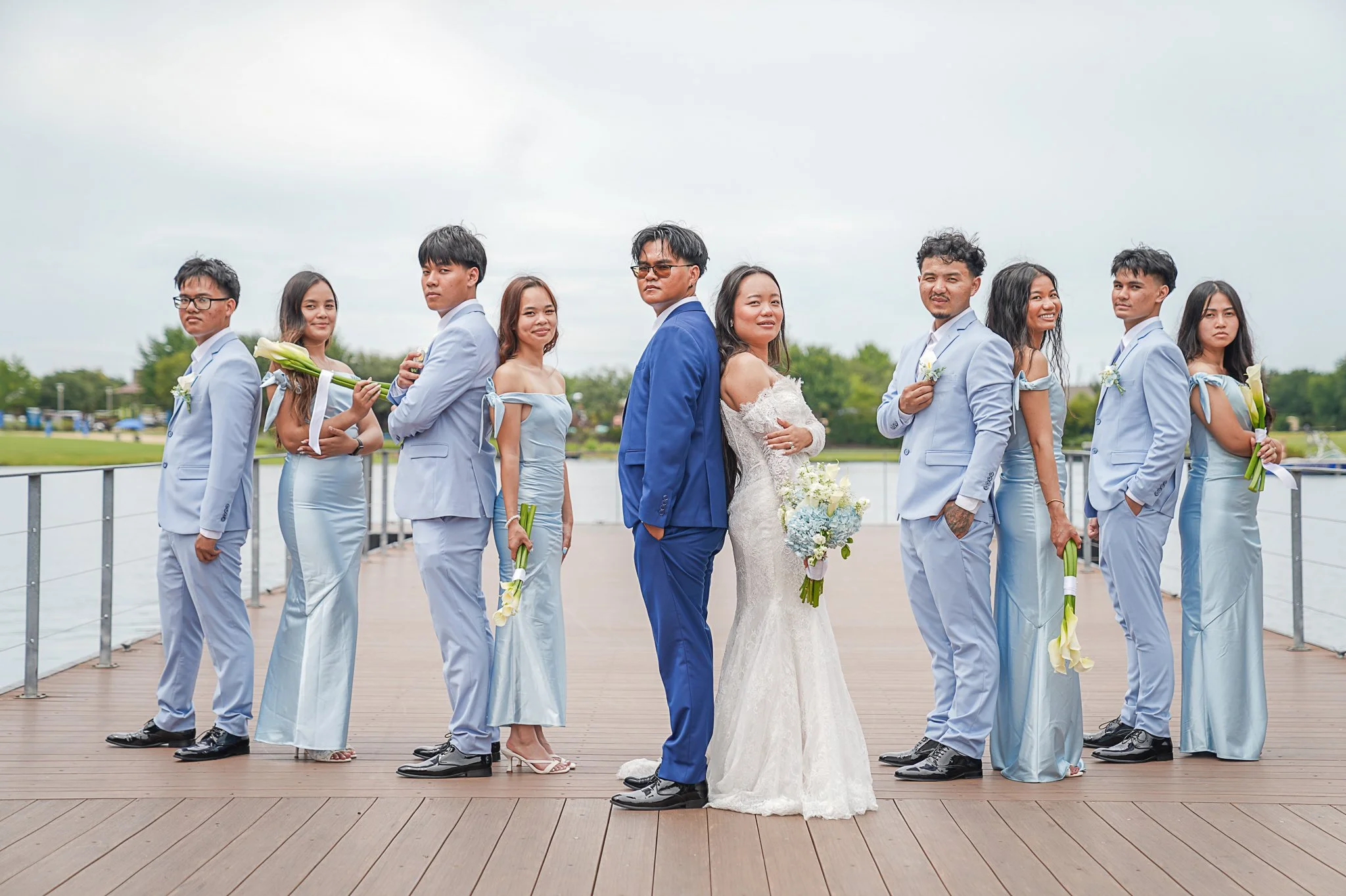 Group of nine people, including bride and groom, dressed in coordinated light blue and white outfits, standing outdoors on a wooden deck near a body of water, with cloudy sky in the background.