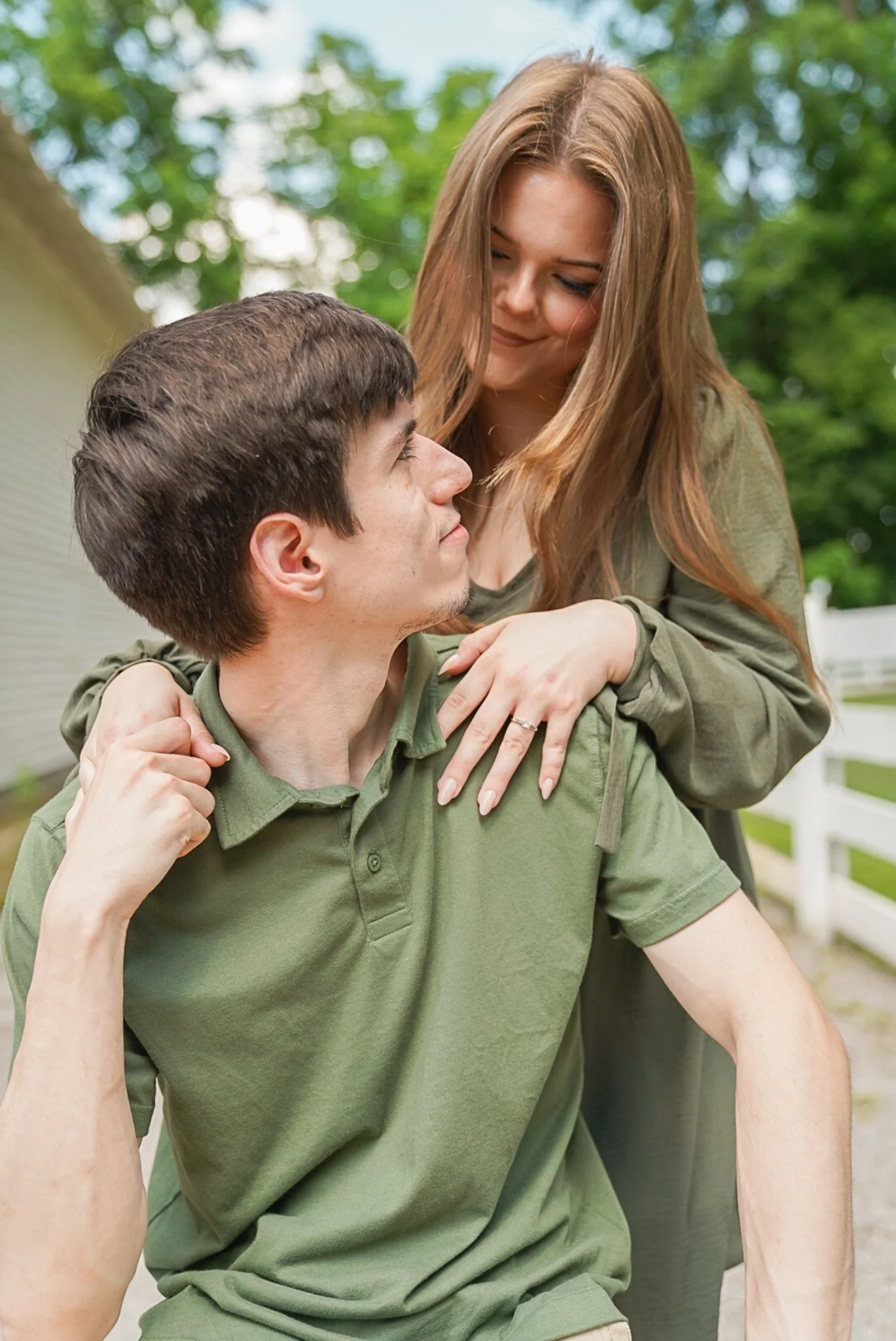 A young man in a green shirt being carried on the shoulders of a young woman, both smiling and looking at each other outdoors with trees in the background.