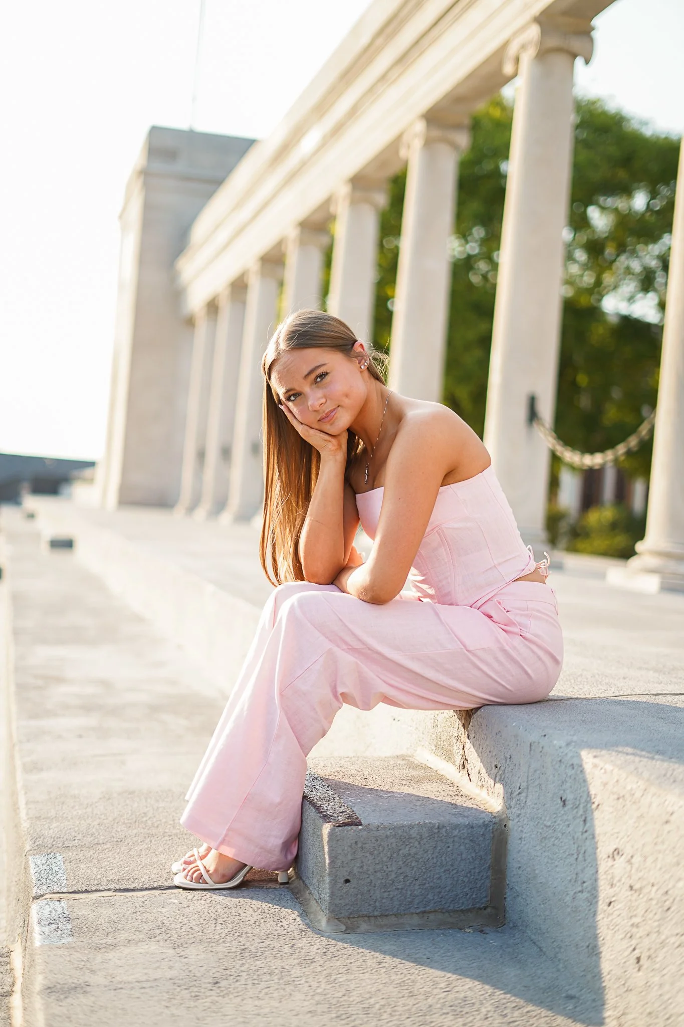 A young woman with long brown hair sitting on a concrete curb in front of a row of white classical columns, outdoors during daytime, smiling gently with her head resting on her hand, dressed in a strapless pink top and wide-legged pink pants.
