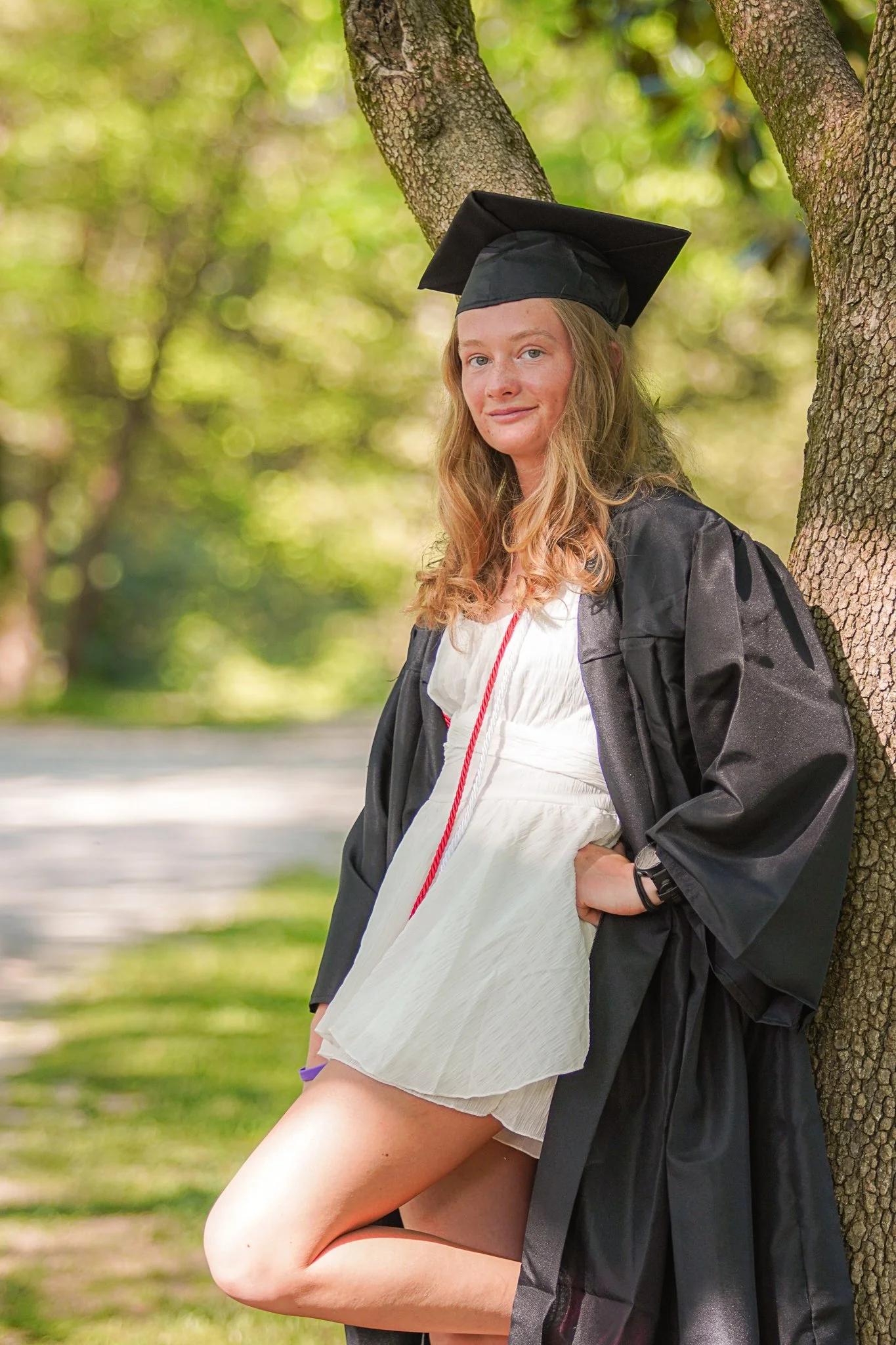 Young woman in a graduation cap and gown leaning against a tree in a park, with sunlight filtering through the trees in the background.