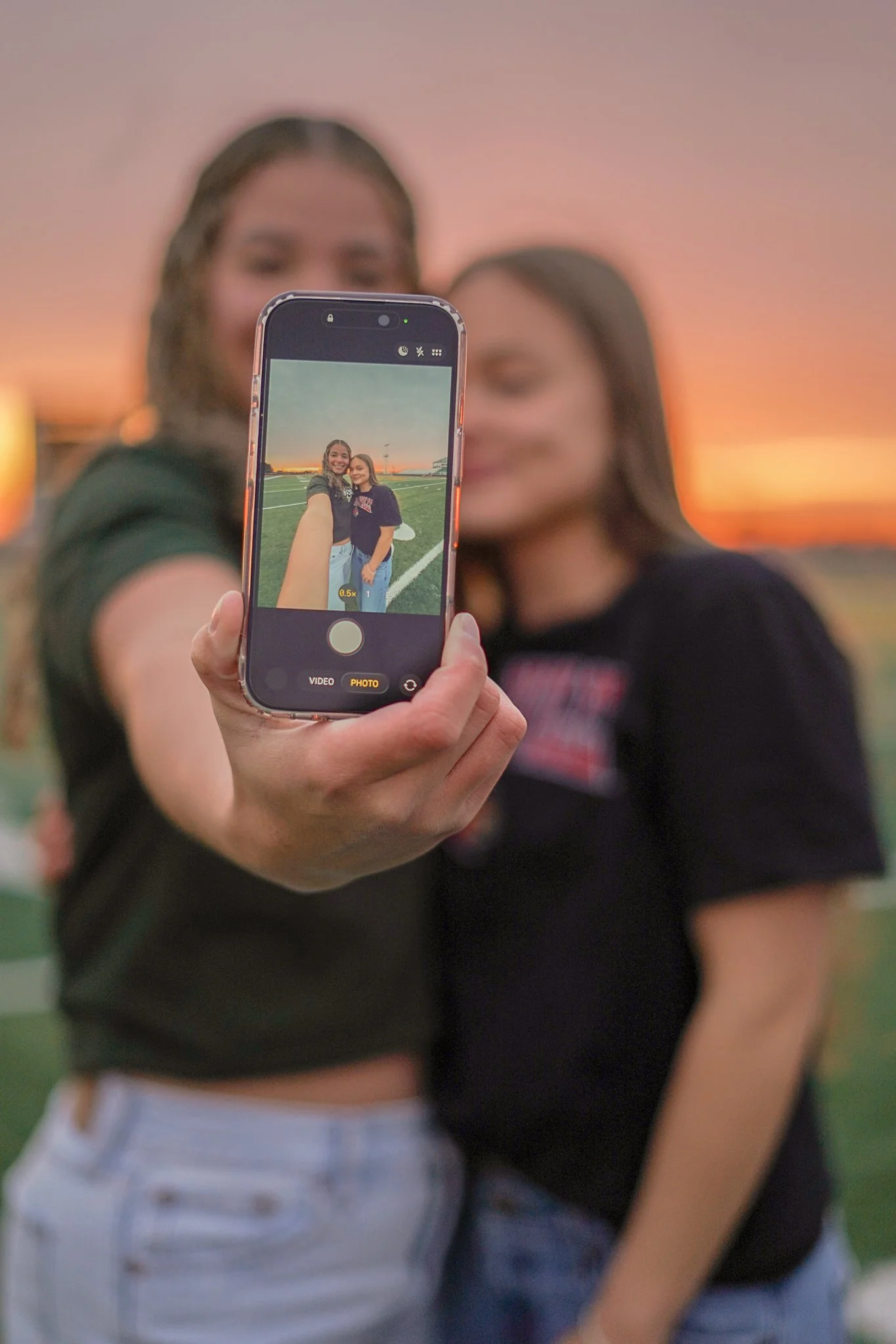 Two young women taking a selfie on a smartphone at sunset, standing on a football field.