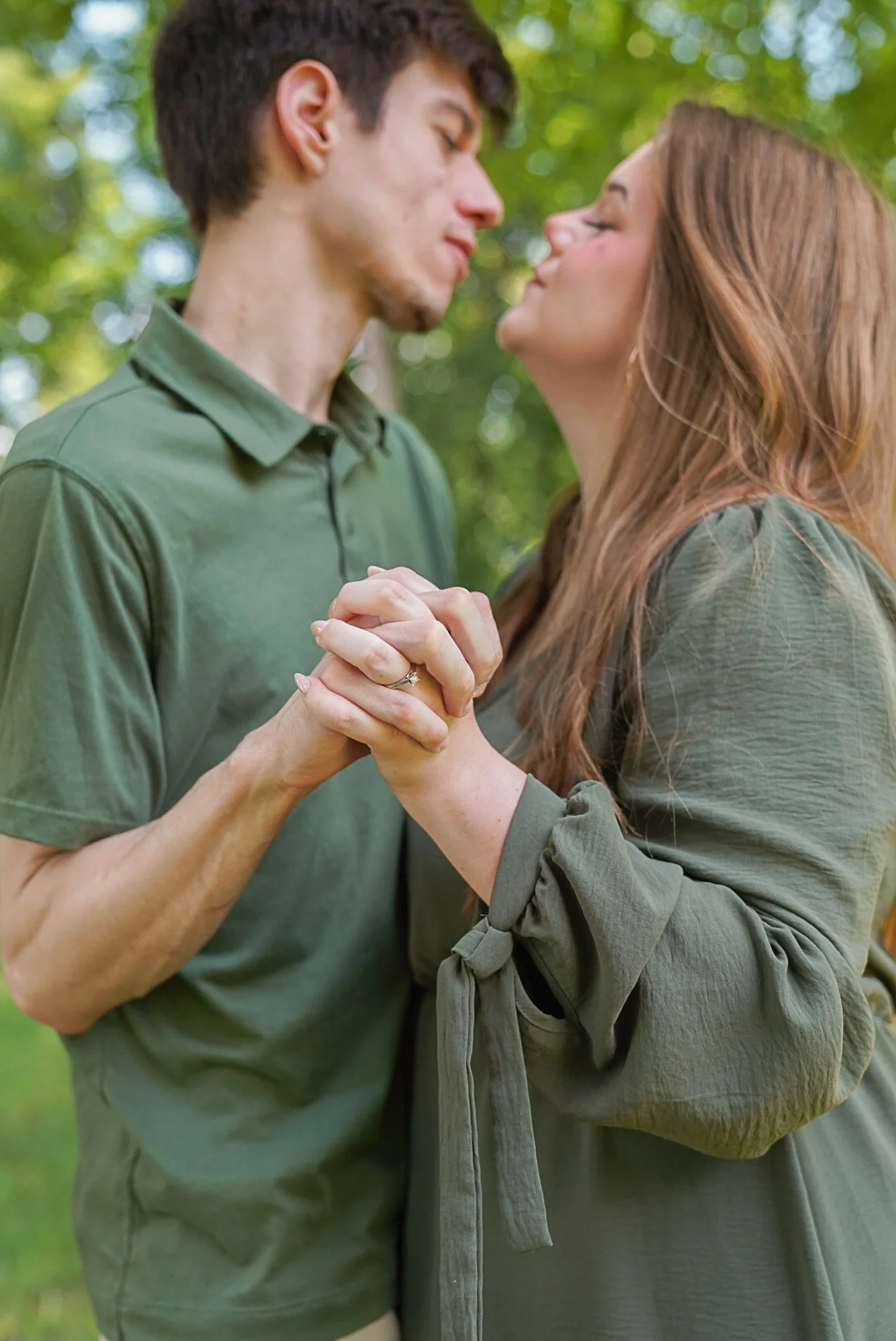 A young couple is dancing outdoors, holding hands and leaning in for a kiss, surrounded by green trees