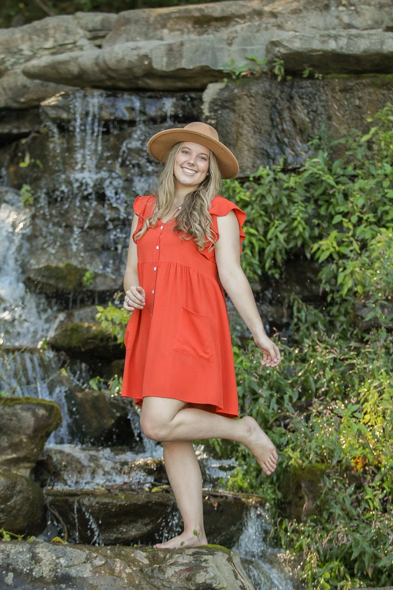 A young woman with long blonde hair smiling and balancing on one foot on a rock in front of a small waterfall, wearing an orange dress and a wide-brimmed tan hat.