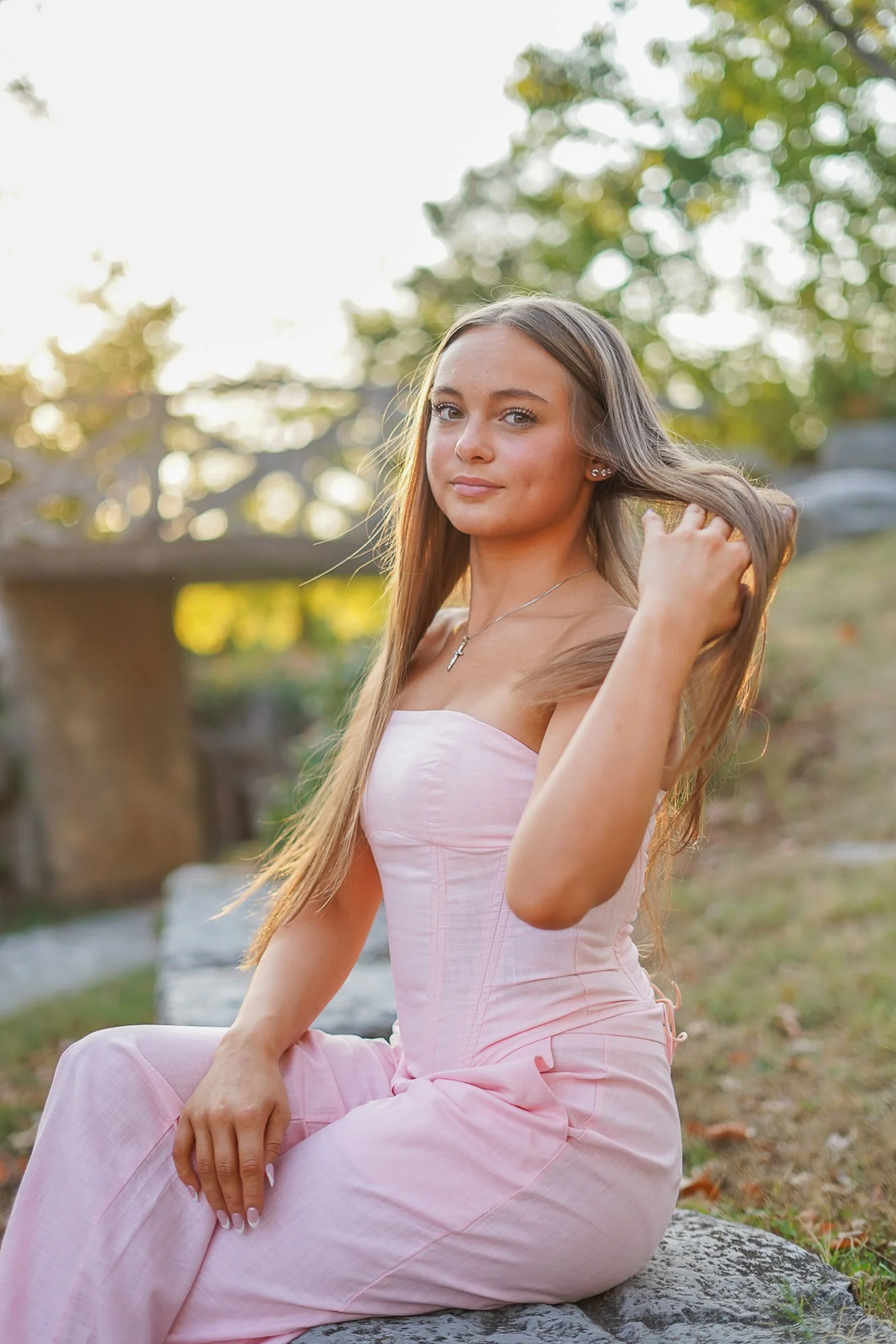 A young woman in a pink dress sitting outdoors on a rock, with trees and sunlight in the background.