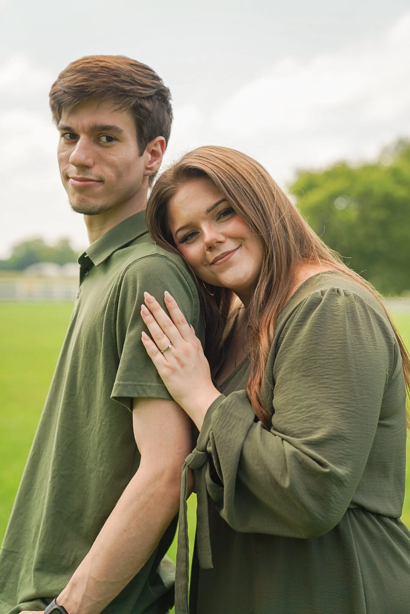 A young man and woman outdoors, standing close together, with the woman leaning her head on the man's shoulder and her hand on his chest. They are both smiling slightly in a green field with trees in the background.