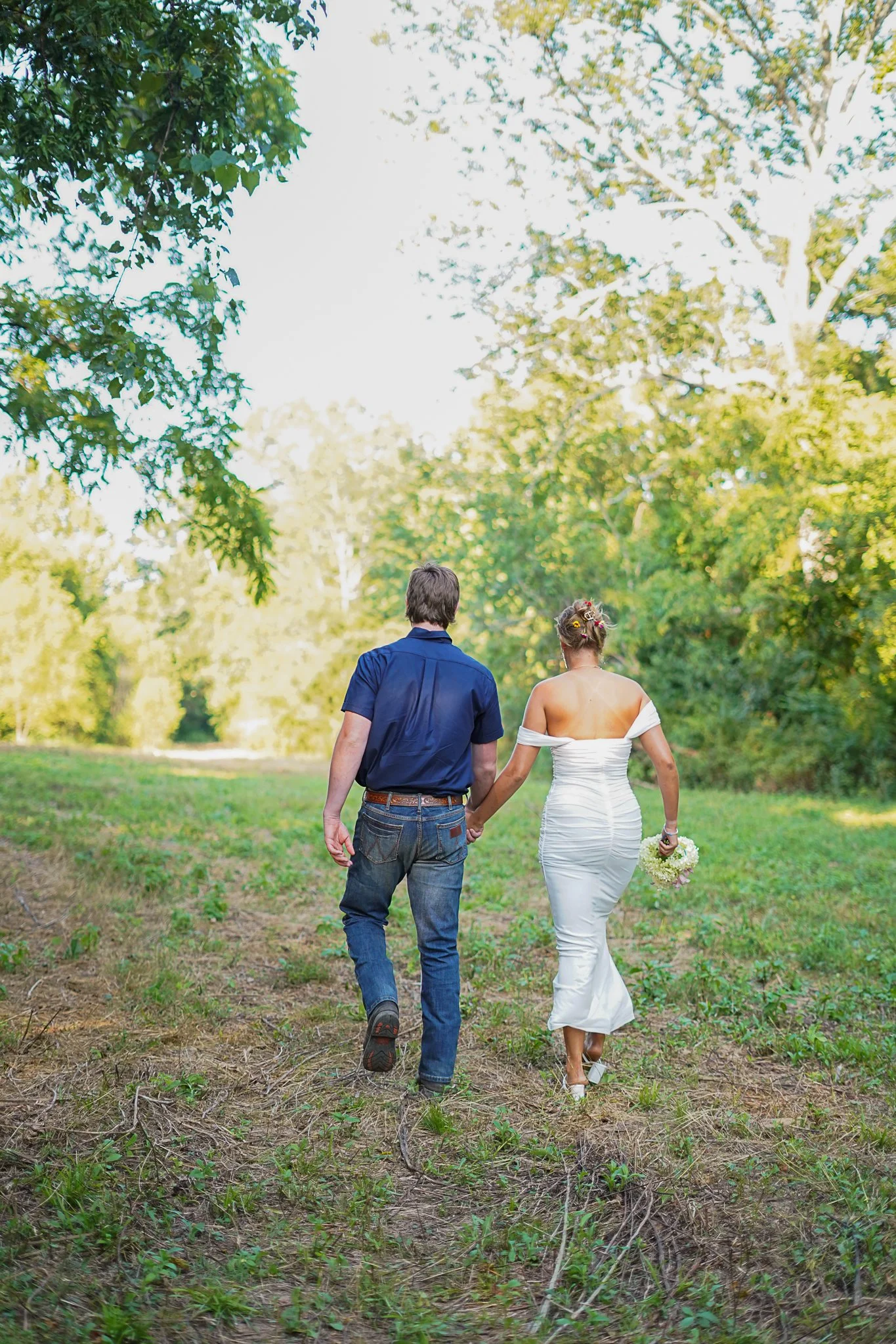 A bride and groom holding hands and walking through a grassy, wooded area during daytime.
