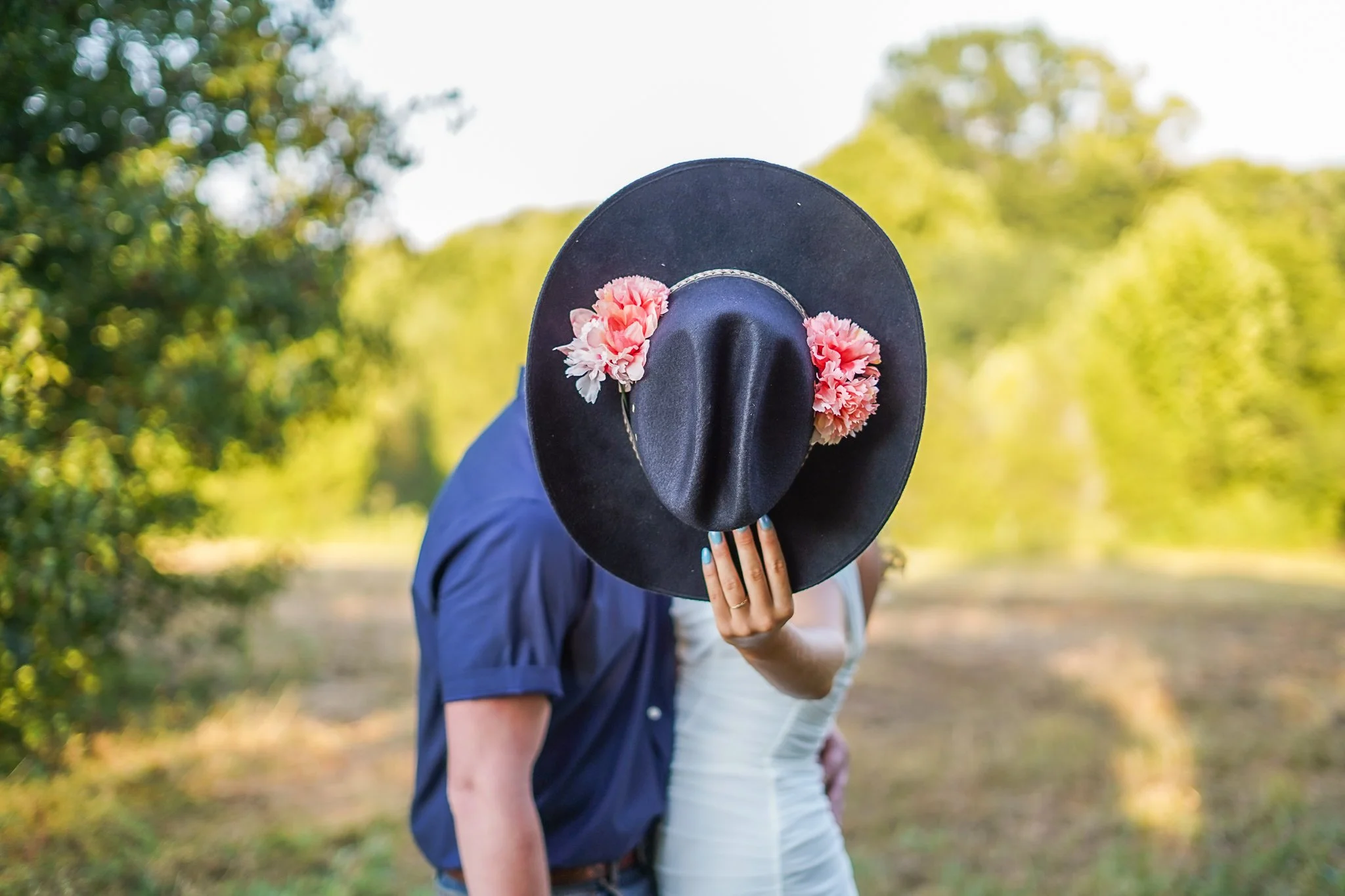 Person holding a black hat with pink flowers in front of their face, outdoors with trees in the background.