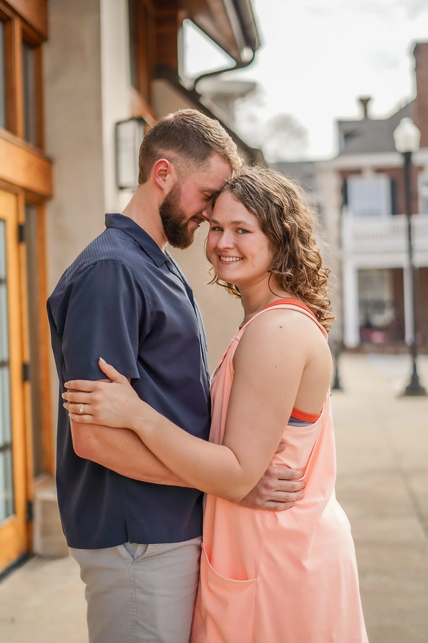 A couple hugging outdoors, smiling and with their foreheads touching. The man is wearing a dark shirt and the woman is wearing a peach-colored dress.