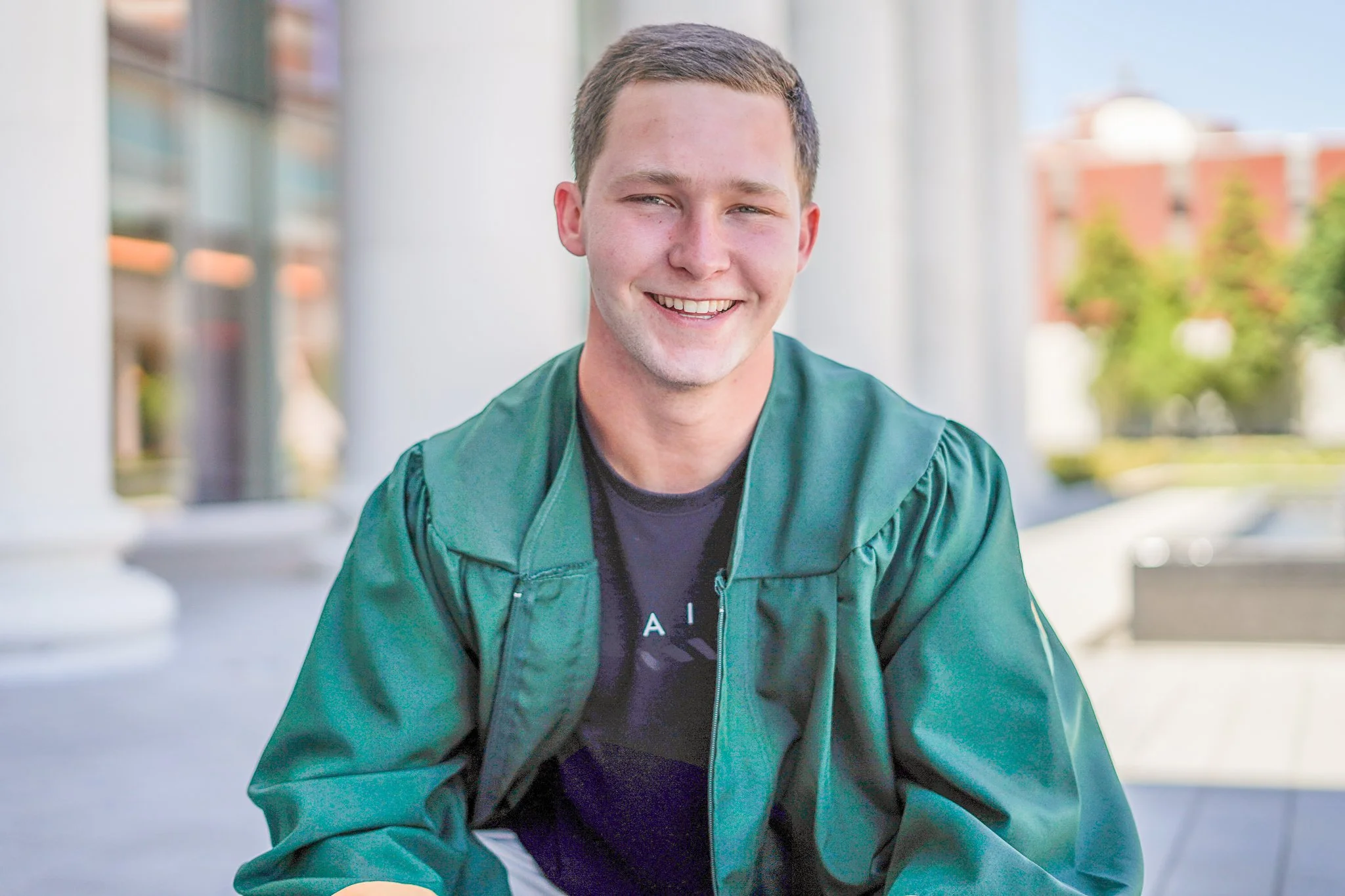 A young man smiling outdoors, wearing a green jacket over a black t-shirt, with a modern building and trees in the background.