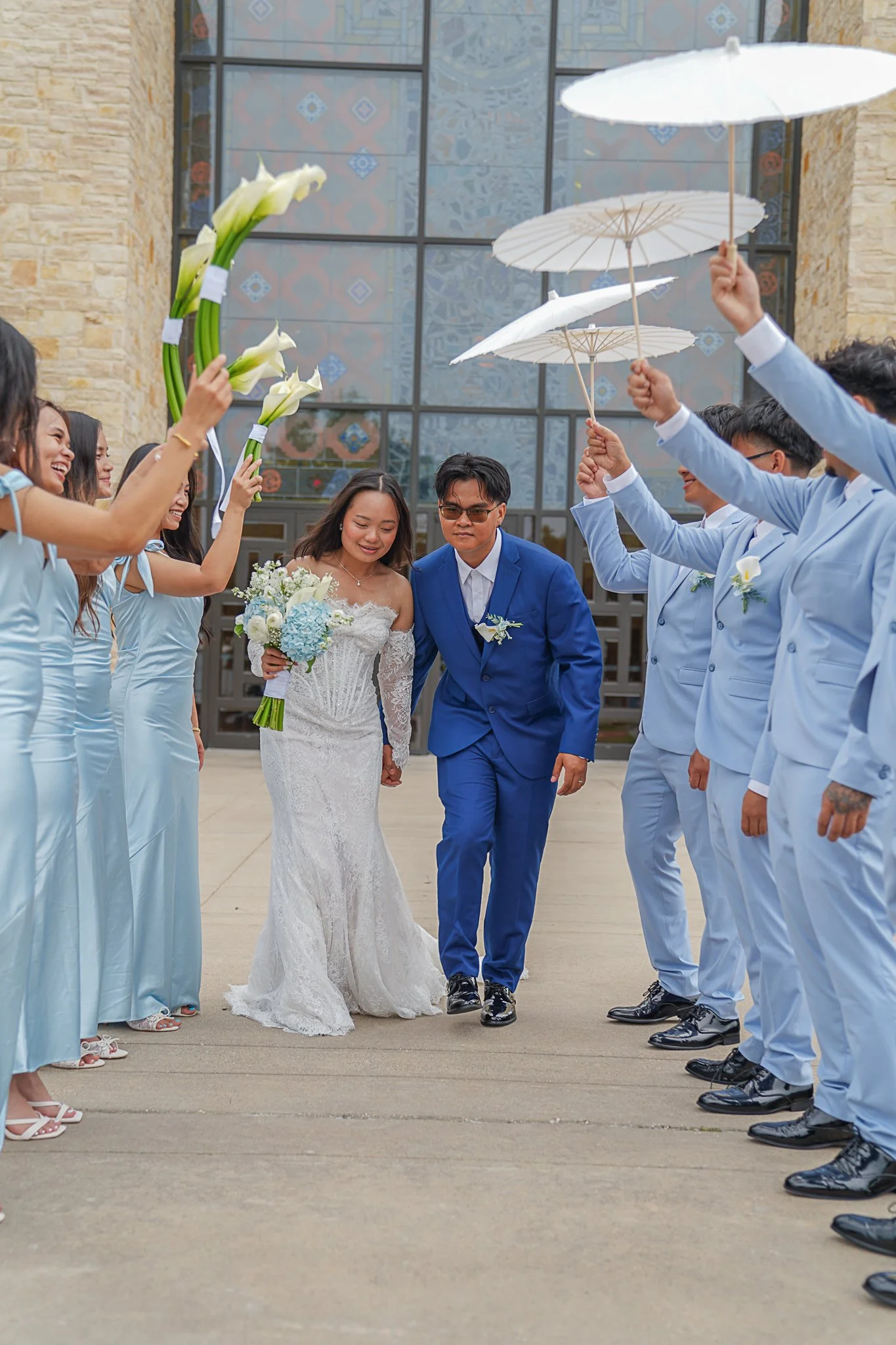 A bride and groom walking together while surrounded by bridesmaids and groomsmen holding umbrellas and bouquets outside a modern building.